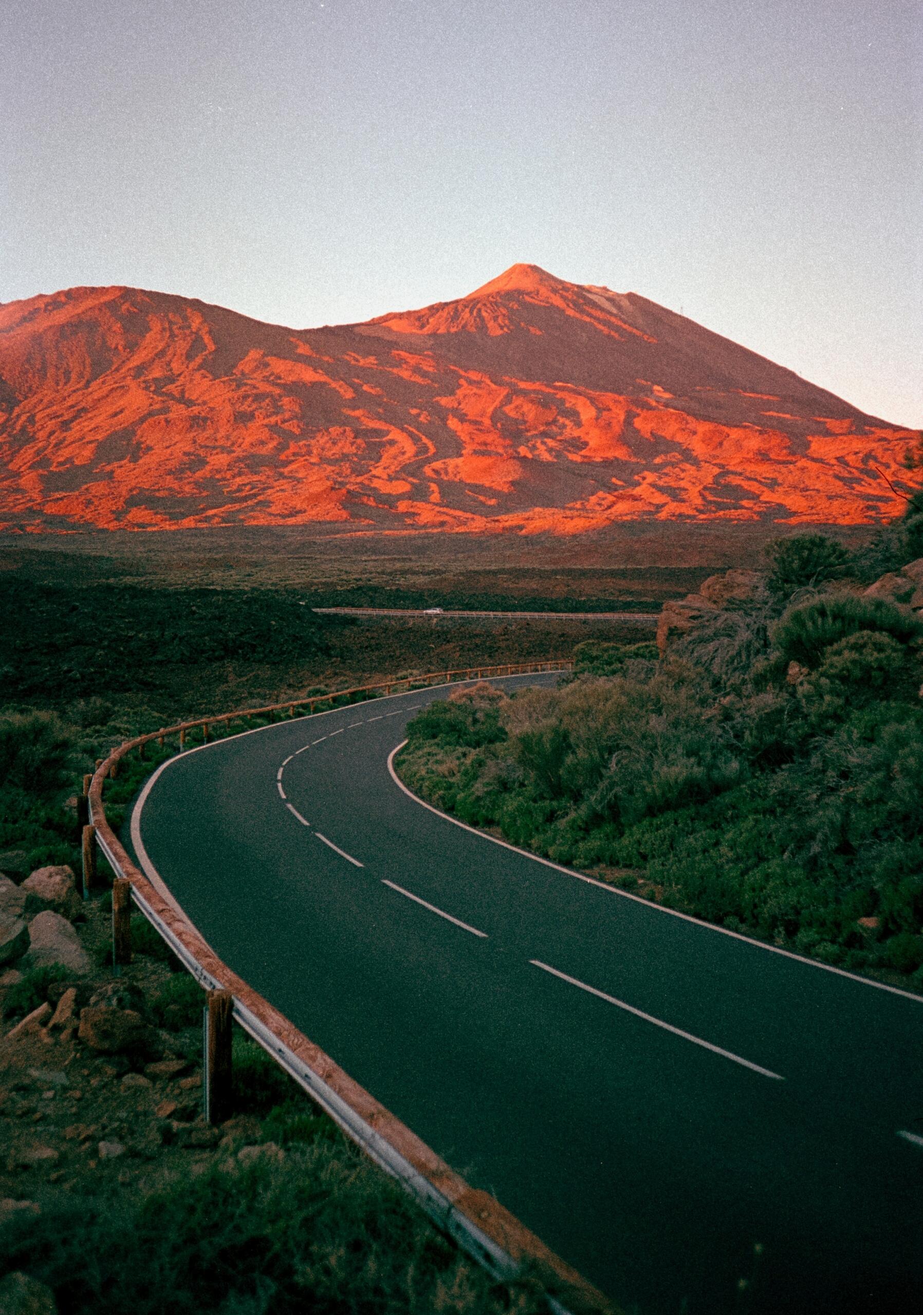 Carretera serpenteante que atraviesa un paisaje pintoresco, con una majestuosa montaña bañada por los cálidos tonos del atardecer al fondo en ternerife