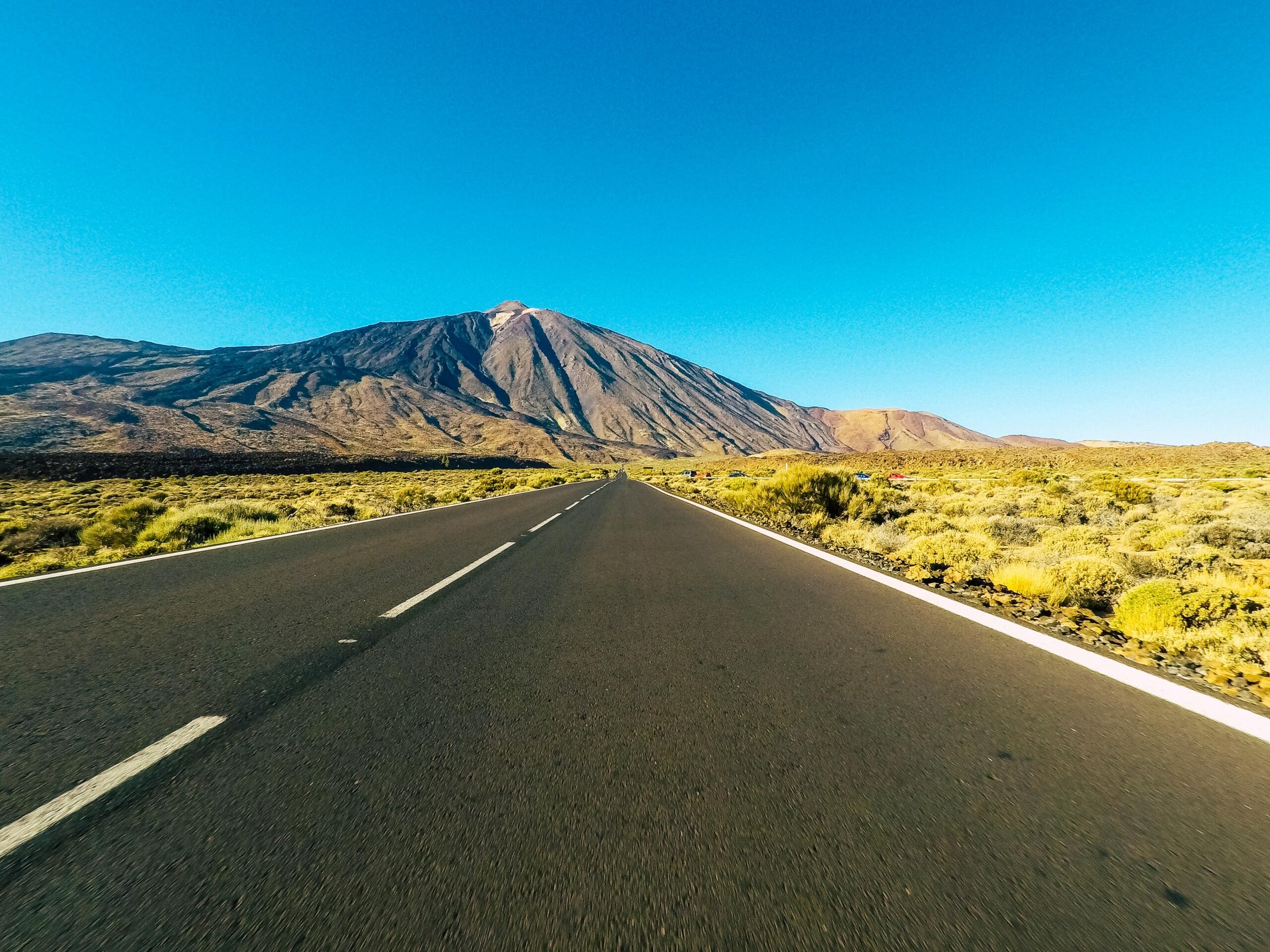 Una carretera recta se extiende hacia el majestuoso volcán Teide, rodeado de paisajes áridos bajo un cielo azul despejado.