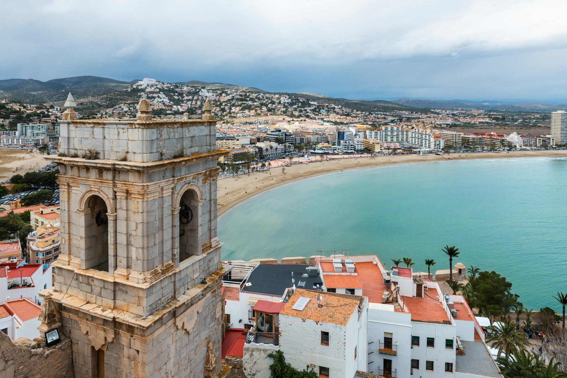 Primer plano de una torre histórica con vistas a una playa y a una ciudad costera, con montañas al fondo bajo un cielo nublado.