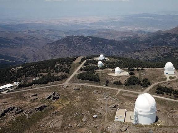 Vista aérea de un paisaje montañoso en el que se aprecian varias cúpulas blancas de telescopios repartidas por un terreno rocoso con vegetación dispersa, bajo un cielo despejado.