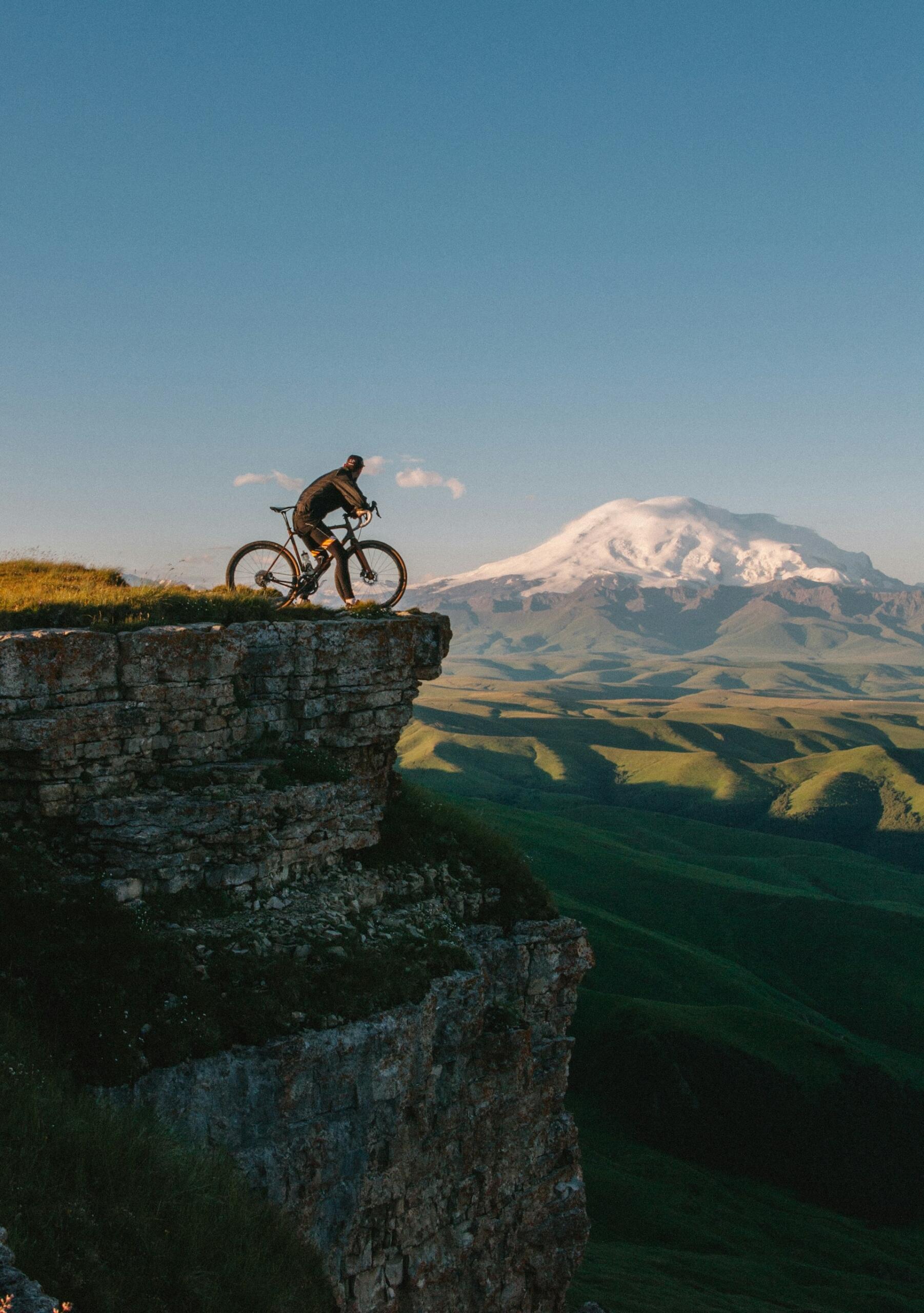 Ciclista en el borde de un acantilado rocoso, con un fondo de colinas verdes y una montaña nevada bajo un cielo azul y despejado, que transmite aventura y emoción.