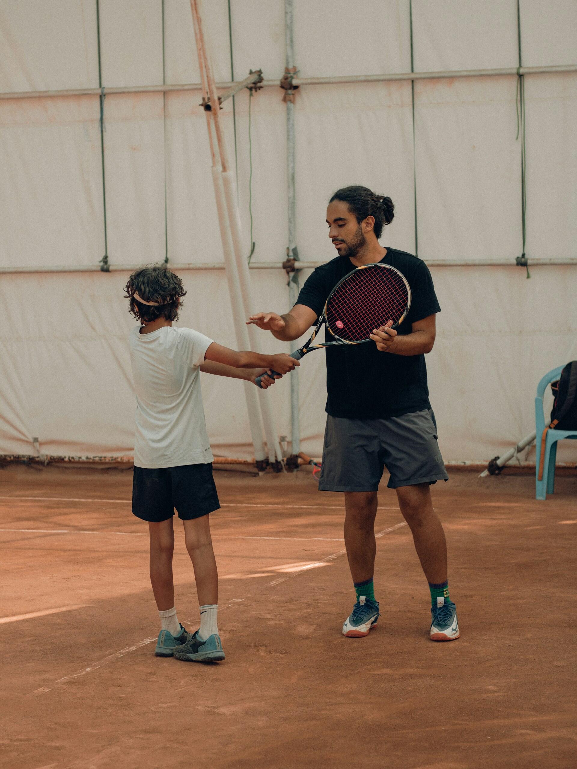 Un entrenador de tenis con camiseta y pantalones cortos negros da instrucciones a un joven jugador vestido de blanco, ajustándole el agarre de la raqueta. Pista cubierta, tono concentrado.