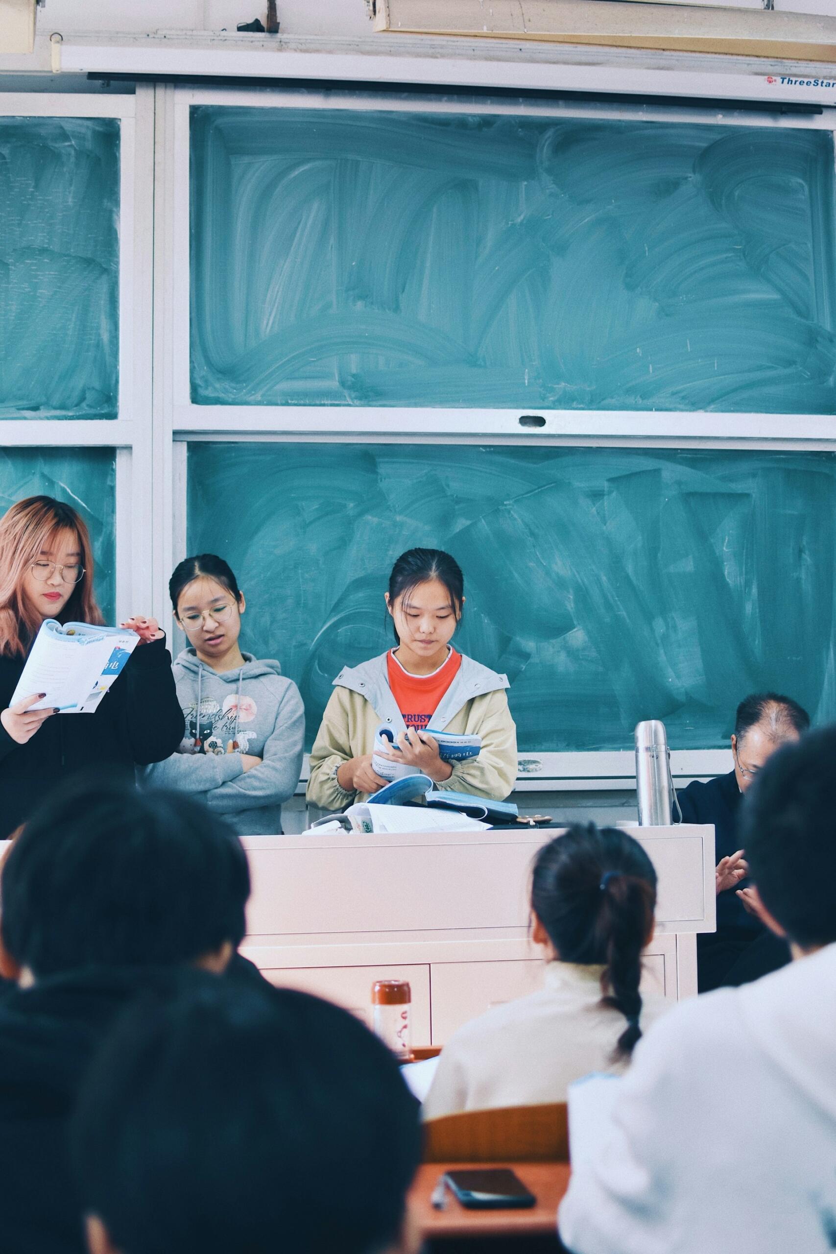 Tres personas están de pie en el estrado de un aula, frente a una pizarra, leyendo libros a un público atento, creando un ambiente de estudio.