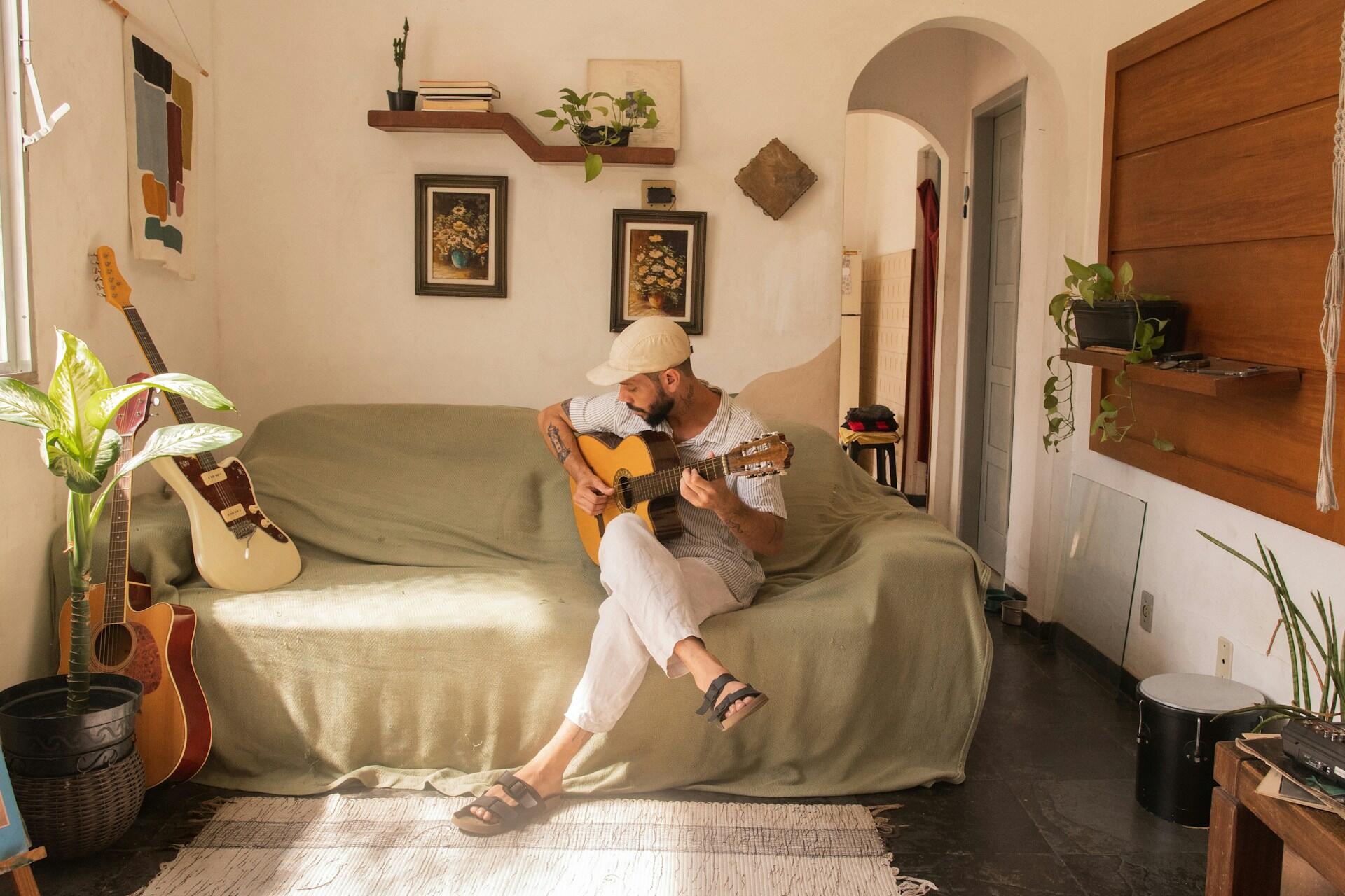 Un músico está sentado en un sofá en una habitación acogedora, tocando una guitarra acústica rodeado de plantas y una decoración artística.