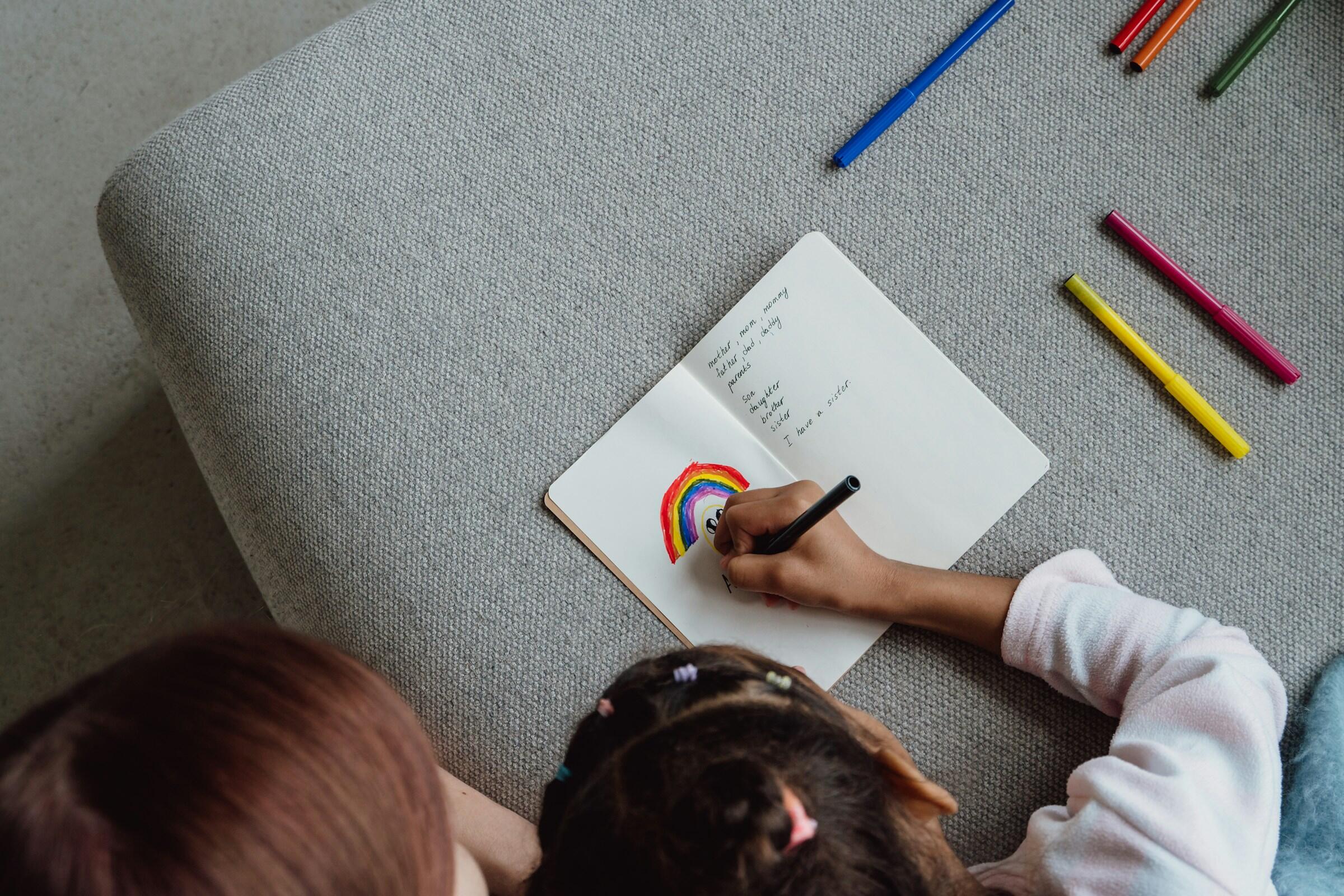 Un niño dibuja un arcoíris colorido en un cuaderno, con rotuladores de colores vivos esparcidos sobre una superficie gris, mostrando su creatividad e imaginación.