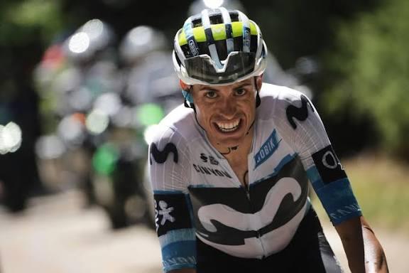 Un ciclista en movimiento, con un maillot azul y blanco y un casco, frunce el ceño con determinación. Al fondo, se ve borrosa una imagen de otros ciclistas y vegetación.