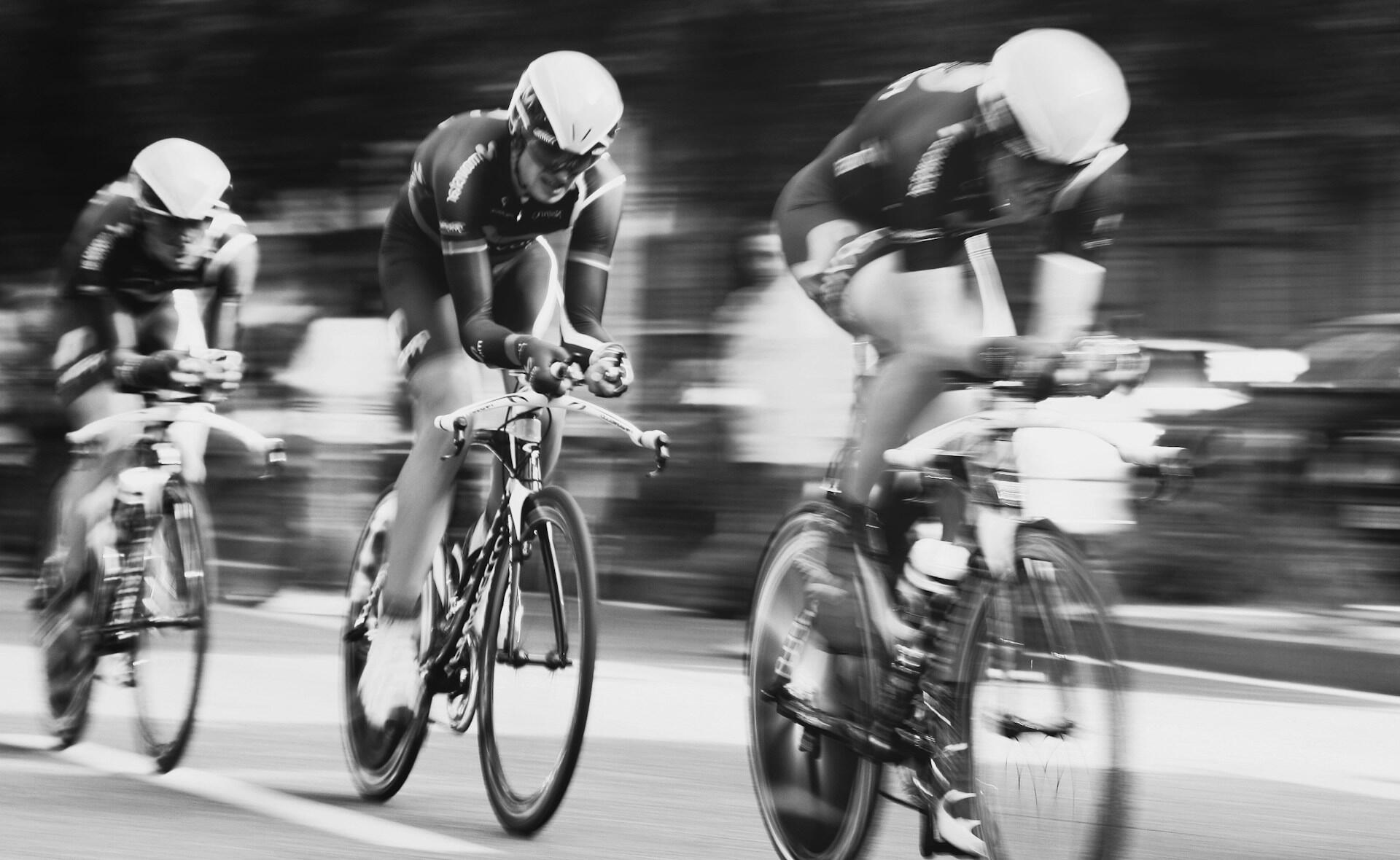 Fotografía en blanco y negro de tres ciclistas de competición compitiendo con cascos aerodinámicos; el movimiento borroso resalta la velocidad y la intensidad en la carretera.