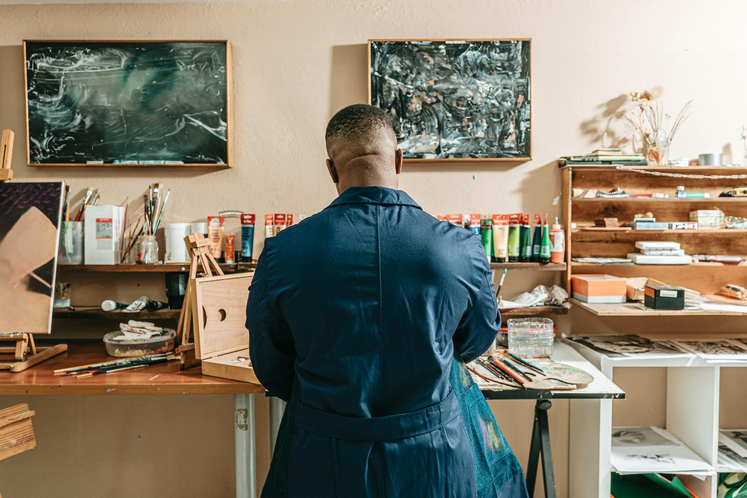 Un artista con bata azul está de pie ante una mesa de estudio desordenada, rodeado de pinturas, pinceles y lienzos inacabados, mirando hacia otro lado.