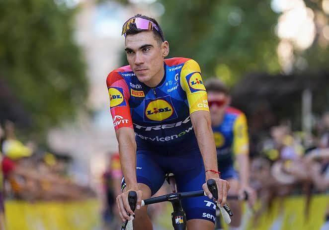 Un ciclista con el colorido uniforme de su equipo pedalea con seguridad, agarrado al manillar. En el fondo, se ven espectadores difuminados y vegetación. Ambiente de concentración y energía.