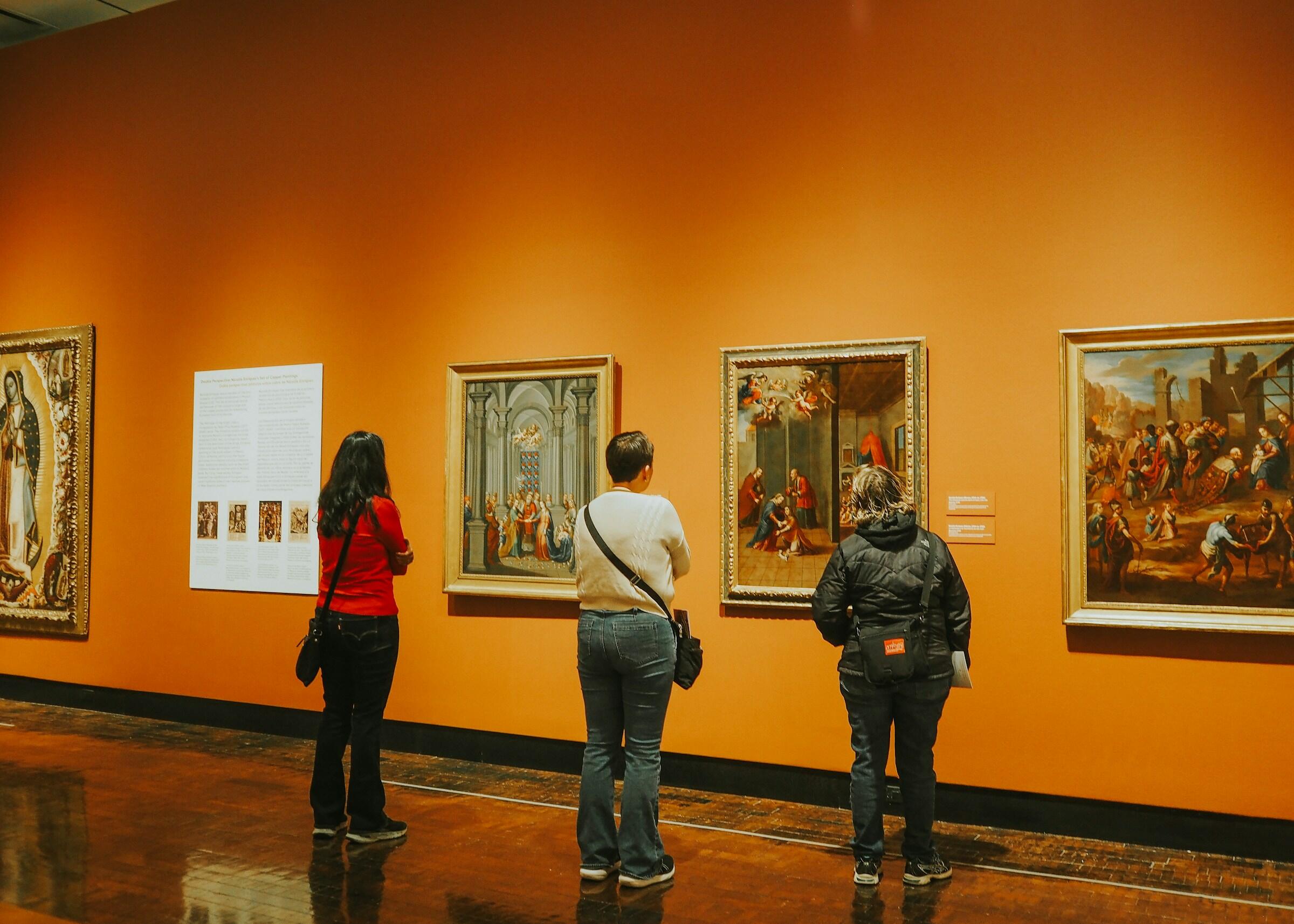 Tres visitantes admiran pinturas clásicas en una pared naranja de un museo, con paneles de texto informativos cerca.