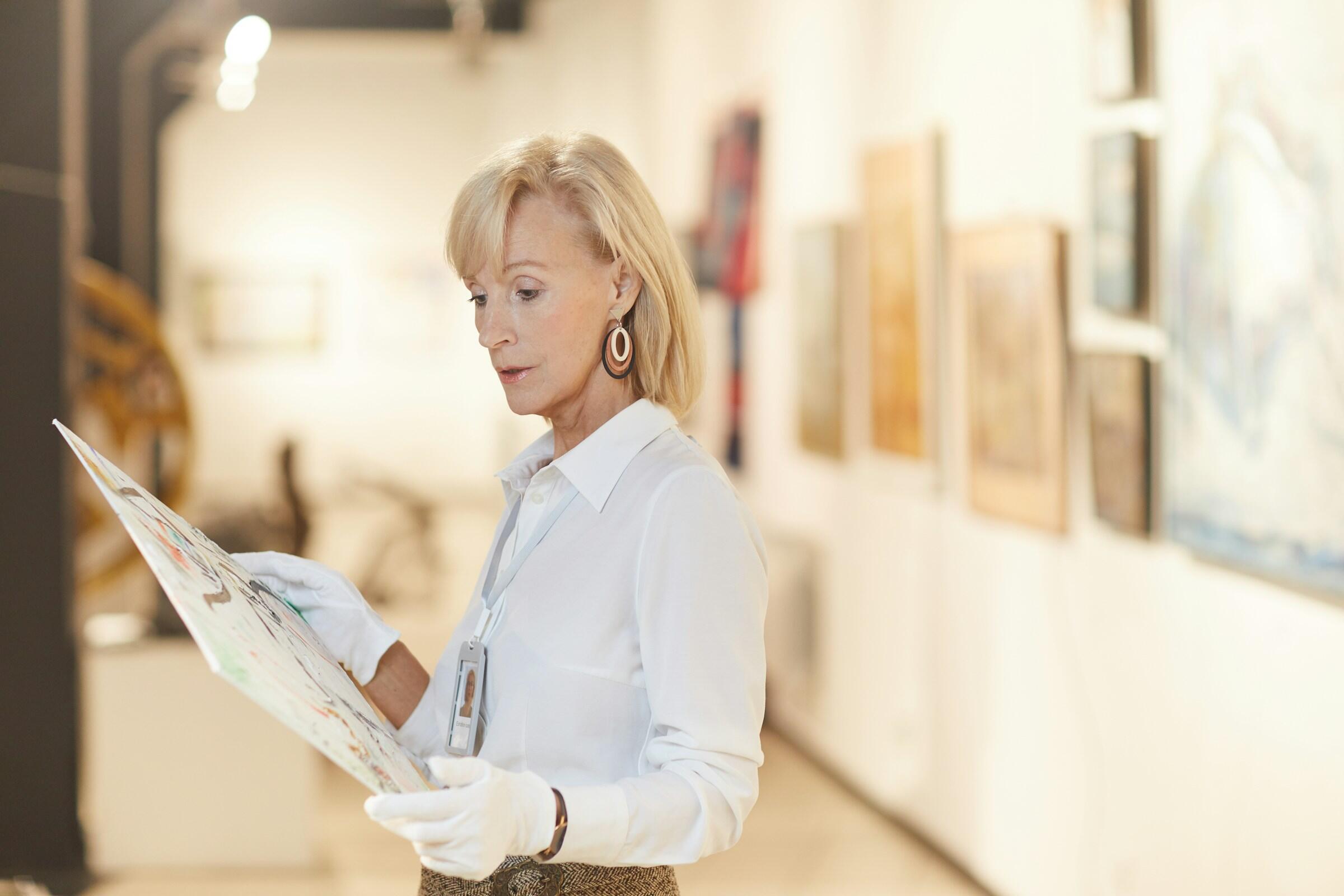 Una mujer en una galería sostiene un folleto de arte, vestida con blusa blanca y guantes, con cuadros de colores expuestos al fondo.