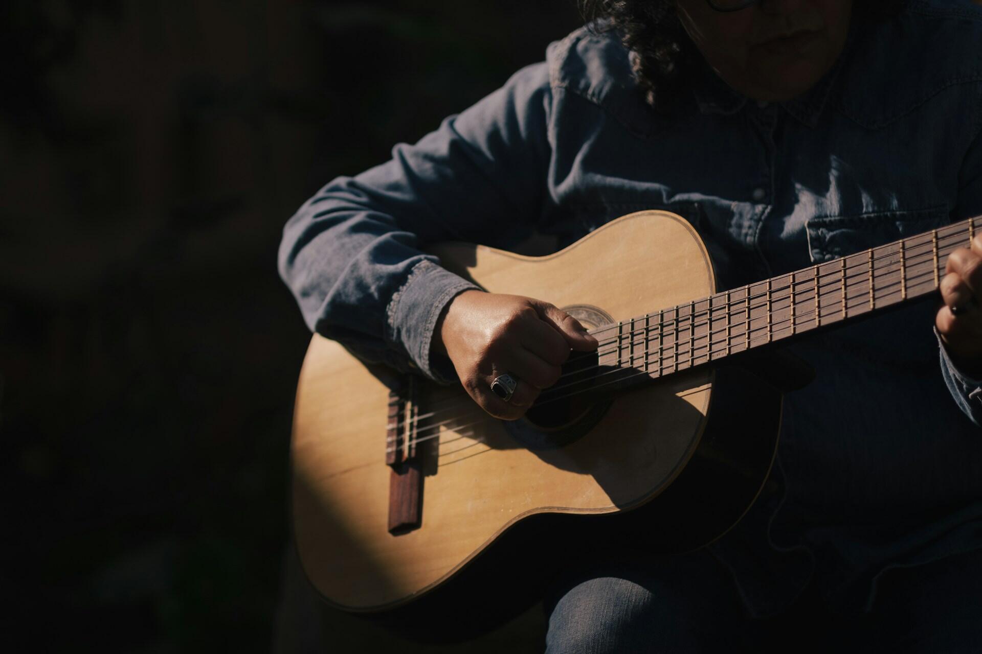 Primer plano de la mano de una persona tocando una guitarra acústica, con un fondo oscuro, resaltando un momento musical de gran concentración.