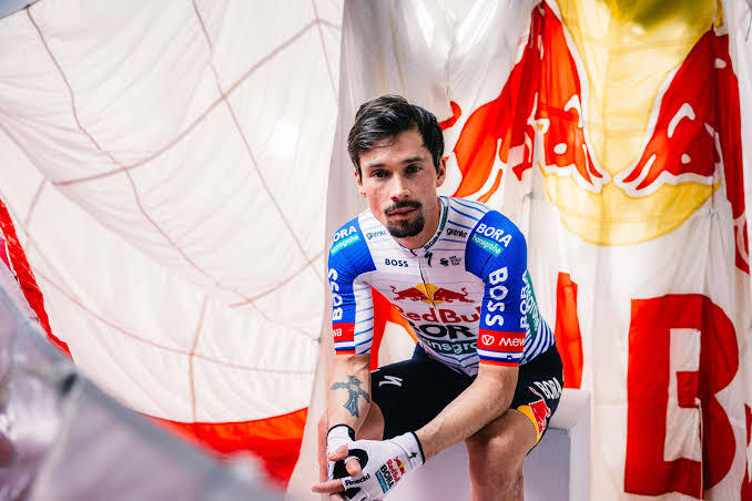 Un ciclista con un maillot azul y blanco con logotipos de patrocinadores se agacha con seguridad, con un gran paracaídas de colores como telón de fondo.