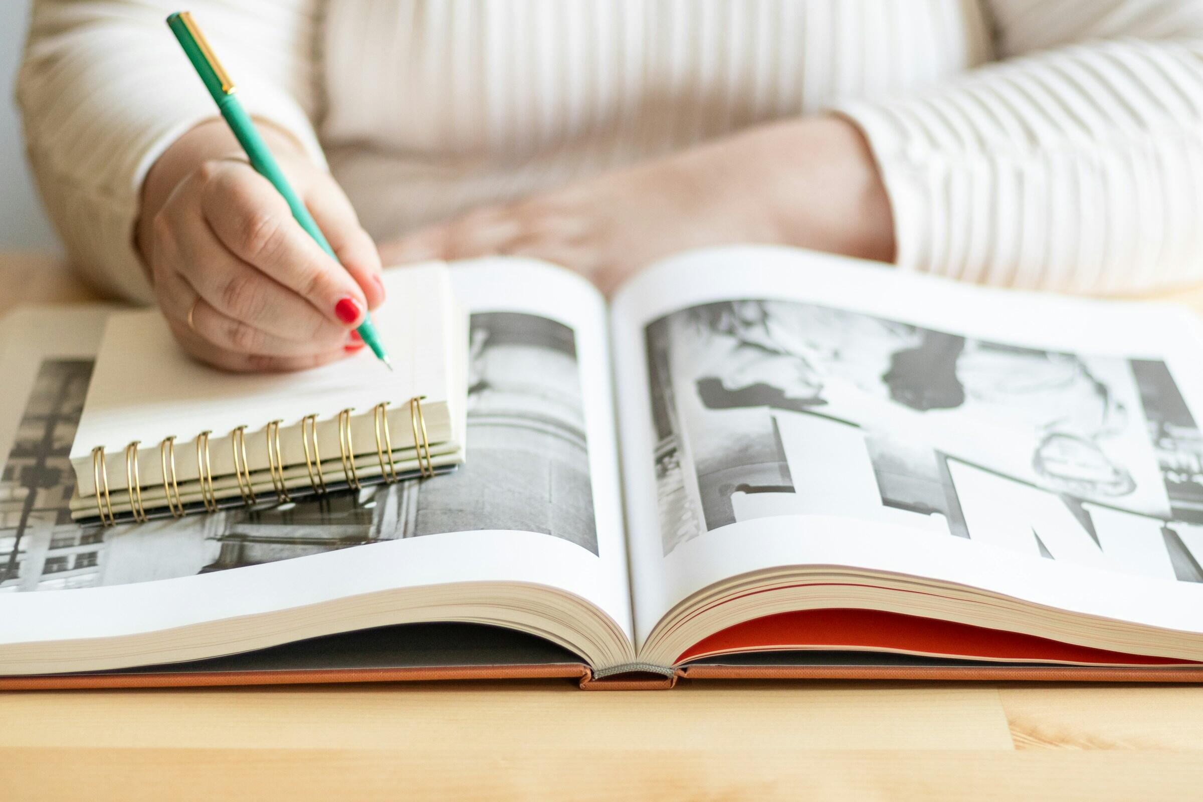 Una persona escribe con un bolígrafo verde en un cuaderno de espiral que tiene junto a una revista abierta.