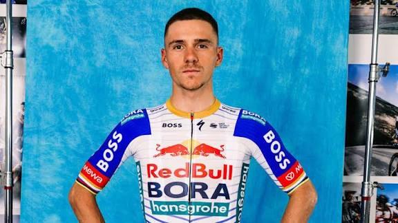 Un ciclista con el maillot blanco y azul del equipo BORA-hansgrohe posa con seguridad ante un fondo azul, en el que se ven patrocinadores como Red Bull y BOSS.