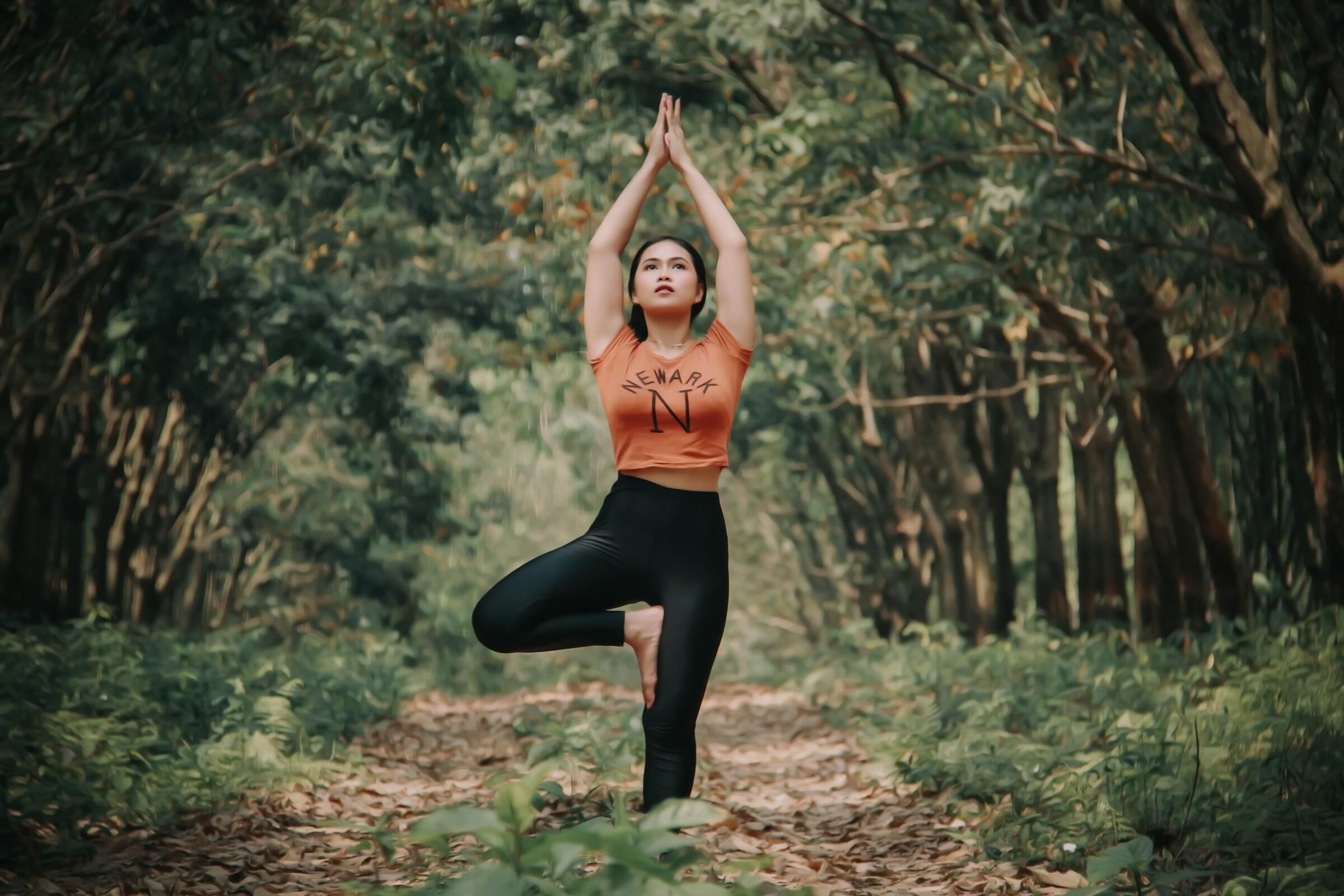 Una persona realiza una postura de yoga en un bosque sereno, rodeada de árboles verdes y hojas caídas en un camino de tierra.