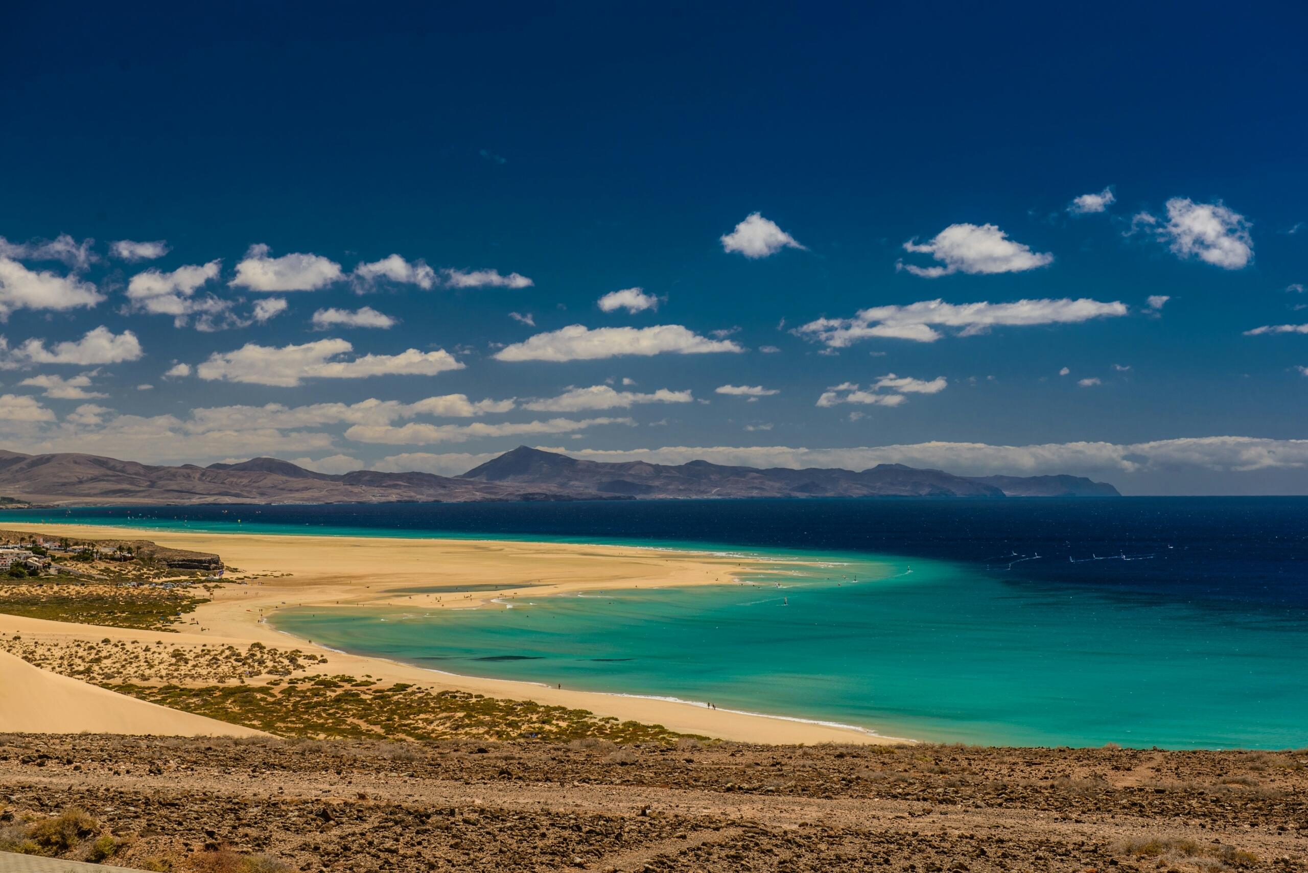 Una vista panorámica de una playa de arena con aguas turquesas bajo un cielo azul brillante, enmarcada por montañas en la distancia.