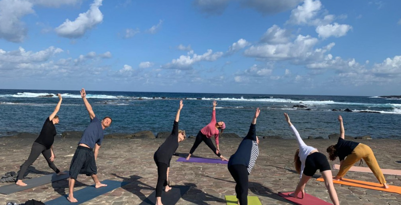 Un grupo de personas practica yoga al aire libre sobre colchonetas de colores, con una vista panorámica del océano y el cielo azul de fondo.