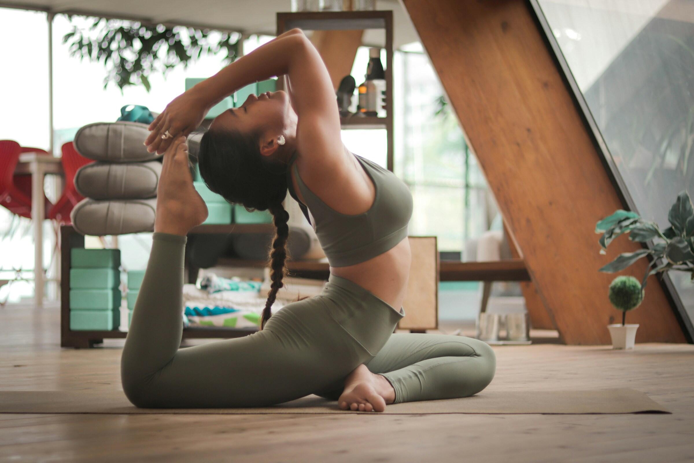 Una persona vestida con un traje verde de yoga realiza una postura flexible sobre una esterilla en un estudio moderno y bien iluminado con plantas y detalles de madera.