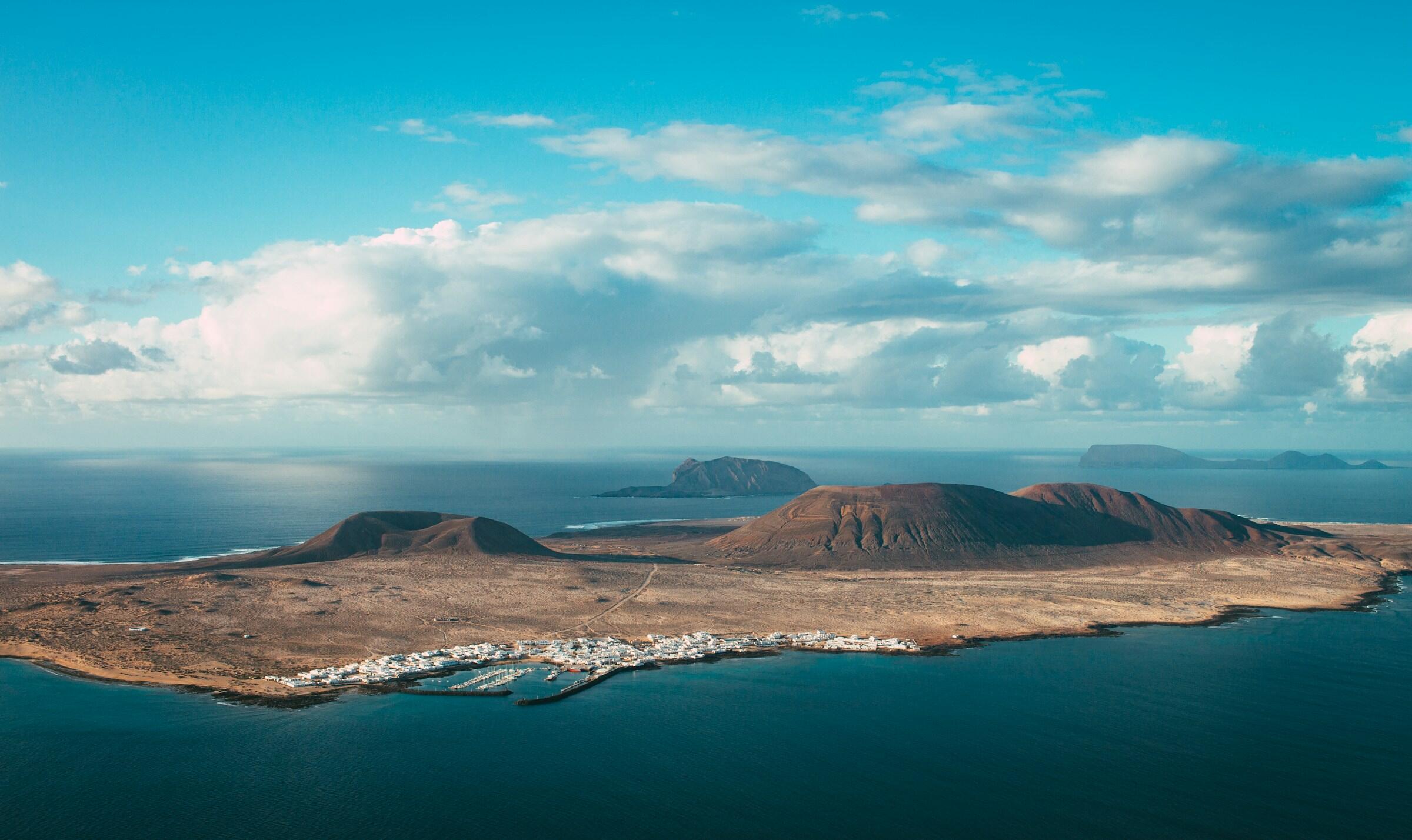 Vista aérea de una escarpada isla volcánica con un pequeño pueblo costero y montañas lejanas bajo un cielo azul con nubes dispersas.