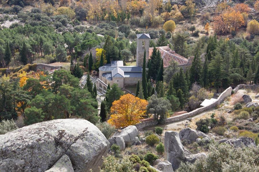 Vista aérea de un sereno monasterio enclavado entre frondosos árboles verdes y follaje otoñal, rodeado de terreno rocoso.