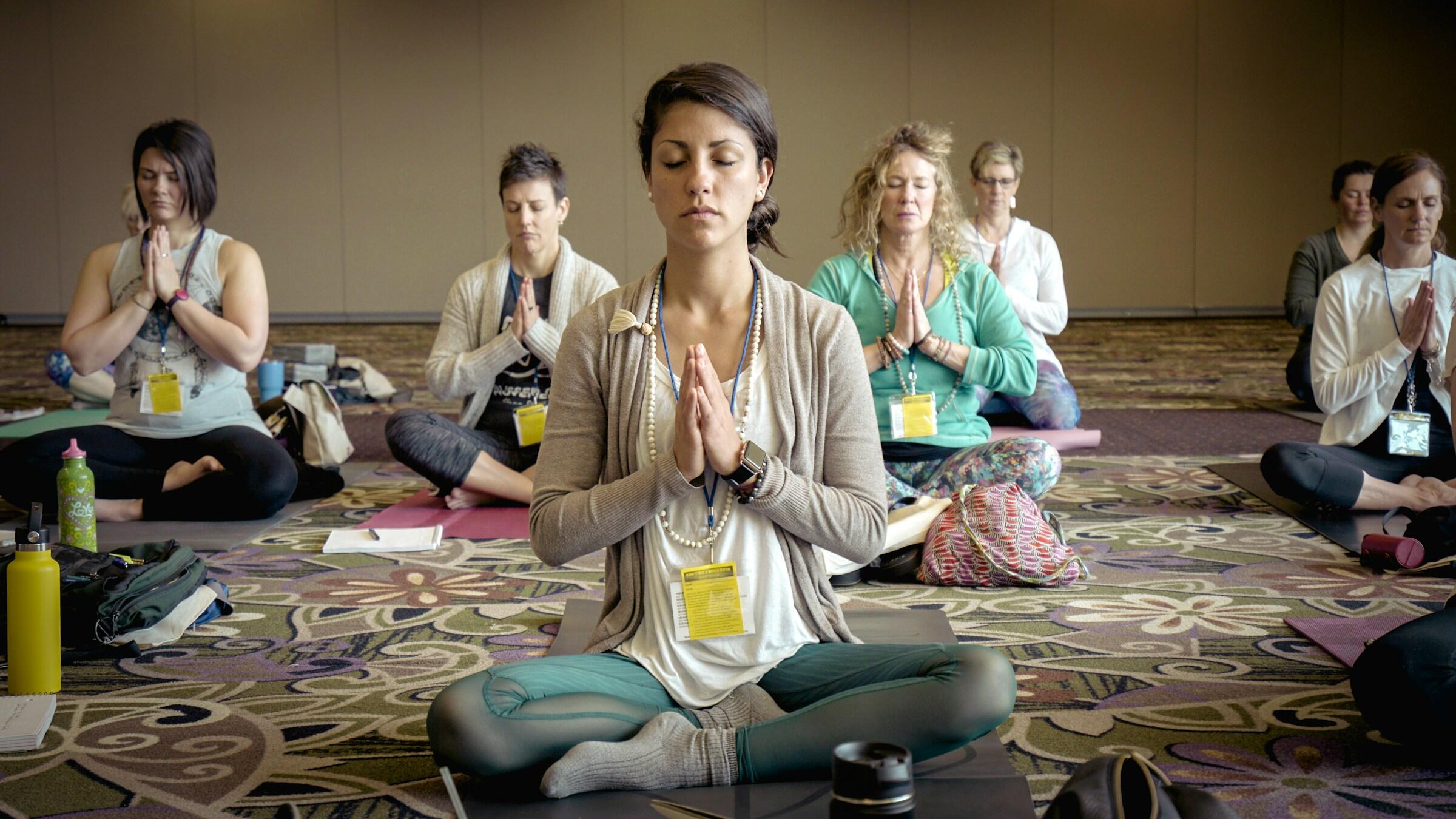 Un grupo de personas practica yoga en una sala espaciosa, sentadas con las piernas cruzadas y las manos en posición de oración, rodeadas de esterillas de yoga y objetos personales.