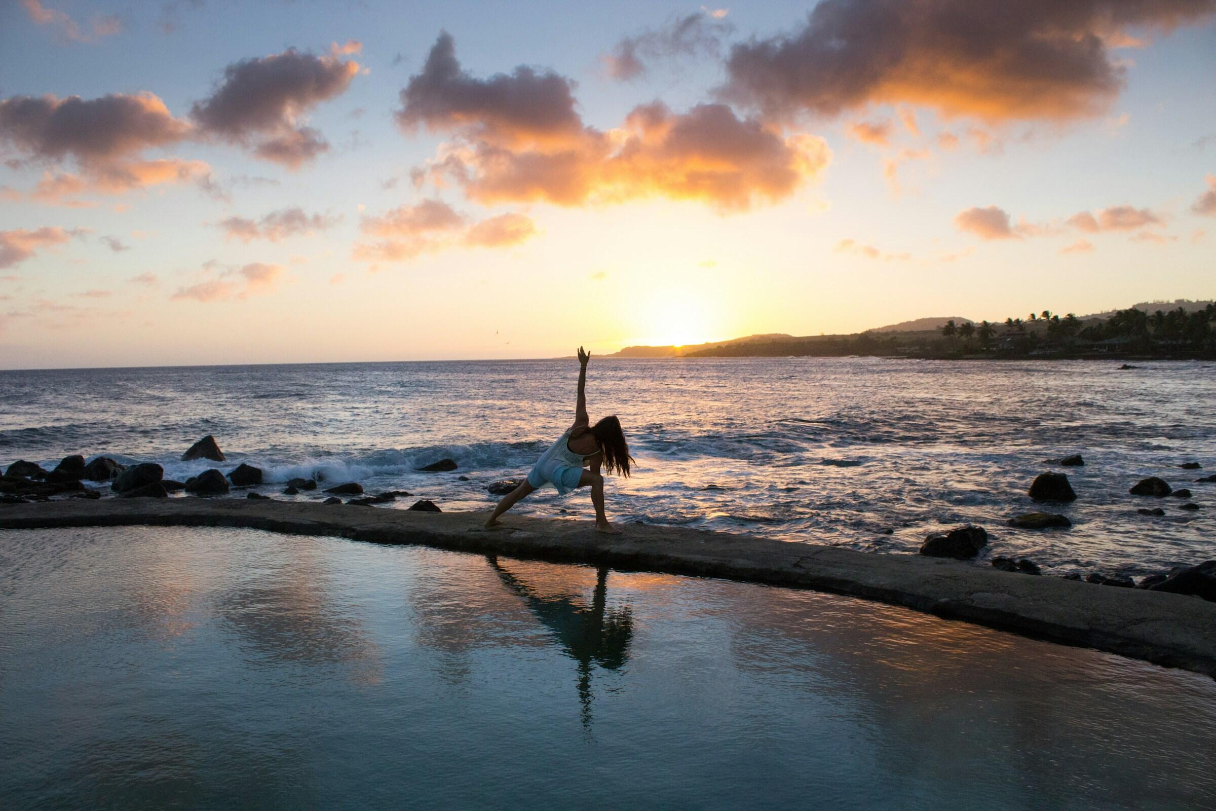 Una mujer practica yoga junto al océano al atardecer, silueteada por nubes de colores que se reflejan en las tranquilas aguas.