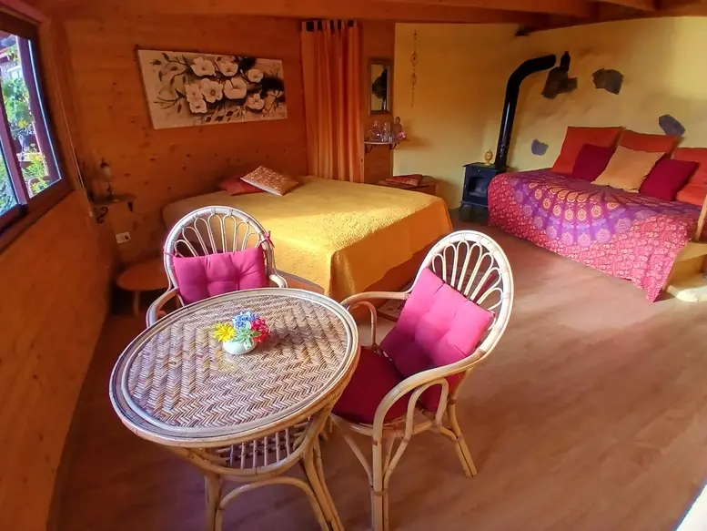 Acogedor interior de cabaña de madera con una cama doble, una colorida zona de estar y una mesita con flores, bañado por una cálida luz.