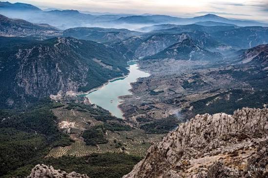 Vista aérea de un tranquilo valle montañoso con un río serpenteante, rodeado de frondosos bosques y acantilados rocosos. Montañas envueltas en niebla al fondo, bajo un cielo nublado.