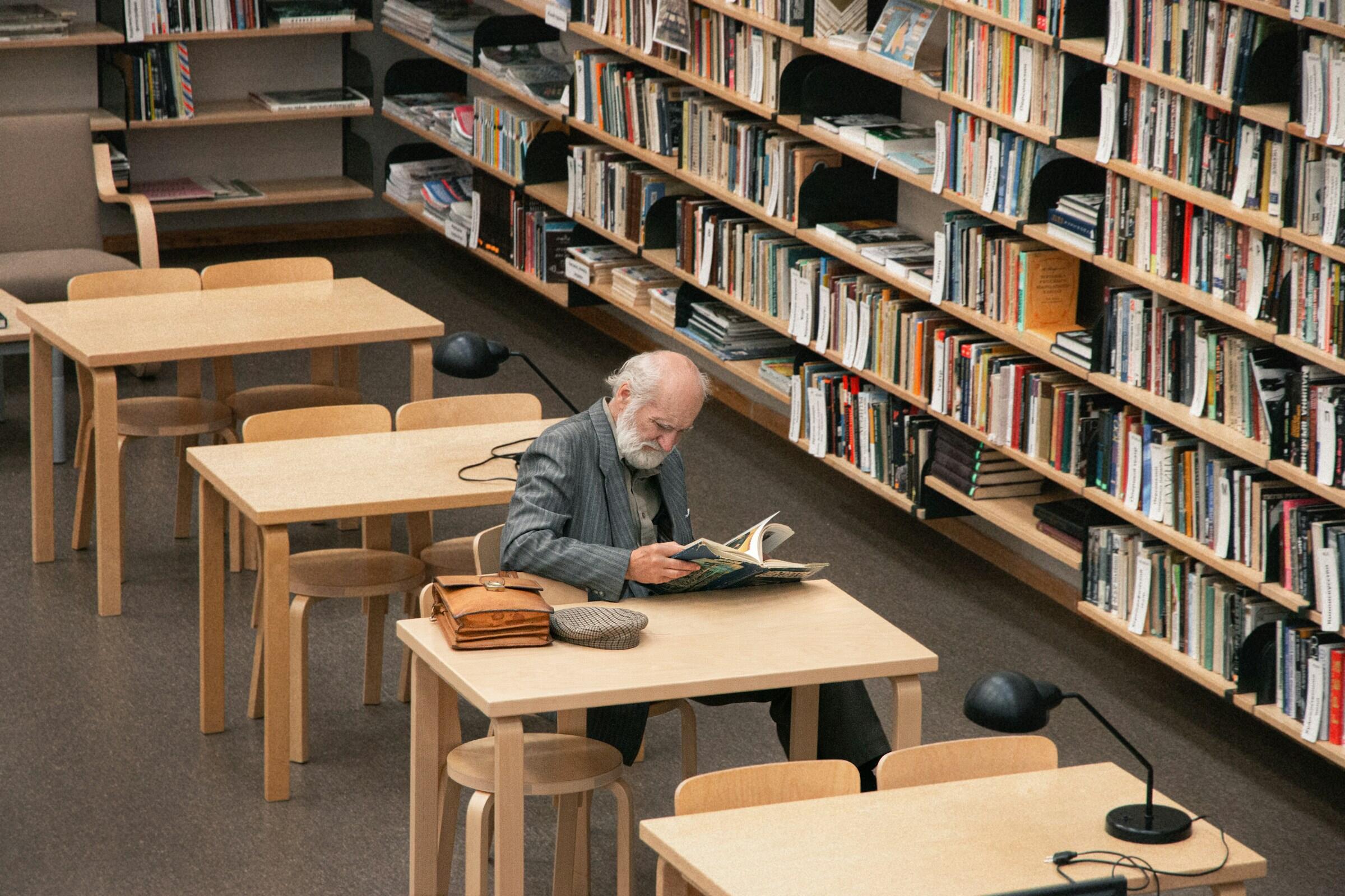 Un anciano leyendo un libro en una mesa de una biblioteca, rodeado de estanterías llenas de libros.