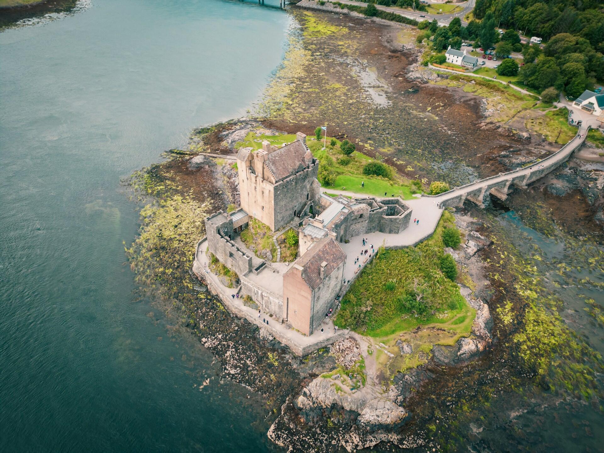 Vista aérea del castillo de Eilean Donan rodeado de agua, con un puente de piedra que lo conecta con la costa y un paisaje pintoresco en las inmediaciones.