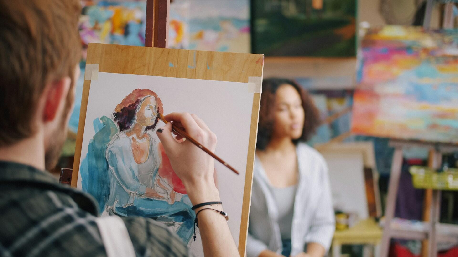 Un artista pinta un colorido retrato de una mujer sentada en un caballete, mientras la modelo se encuentra sentada cerca de él en un estudio de arte luminoso y creativo.