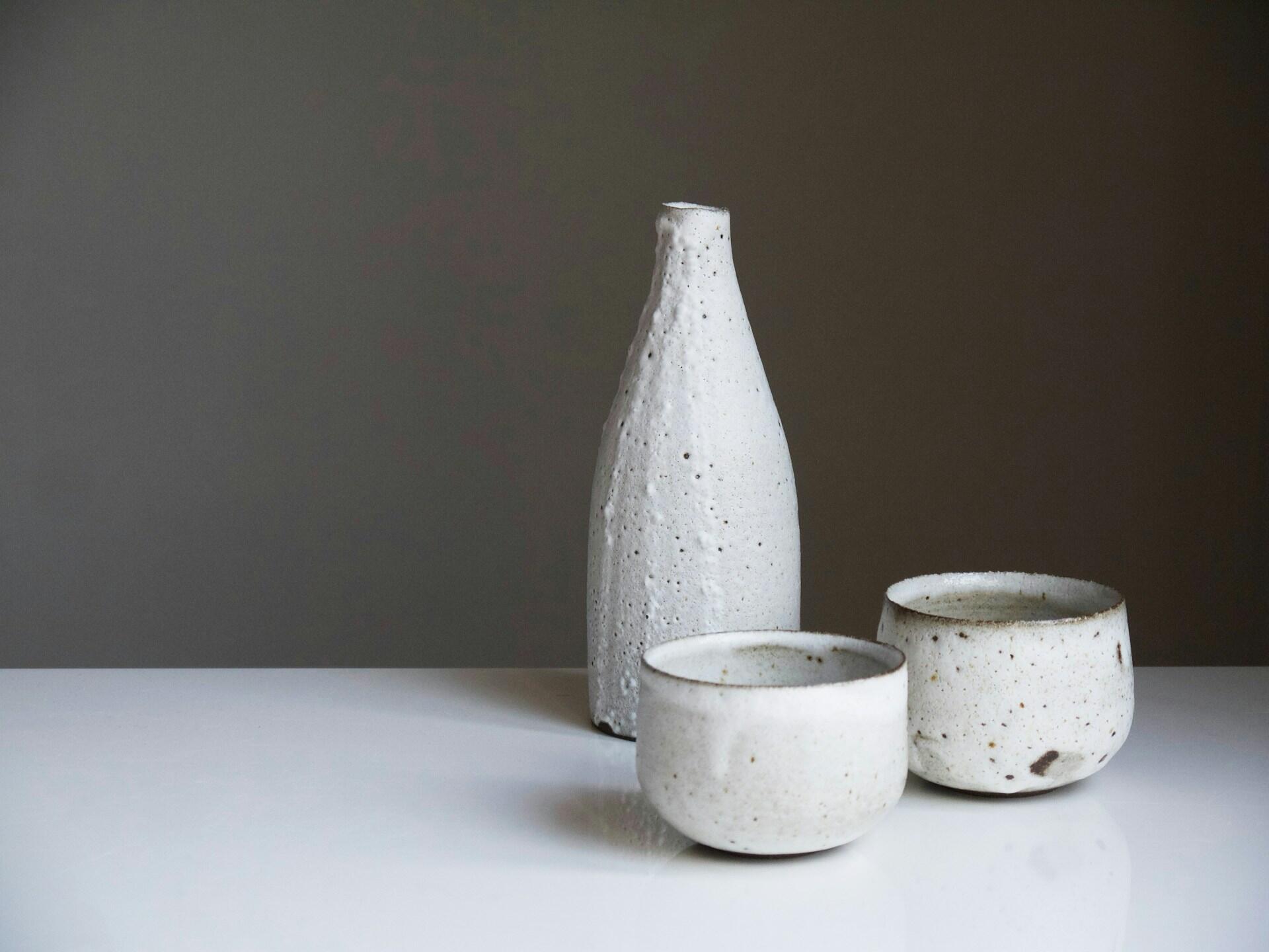 Un jarrón de cerámica blanca texturizada y dos cuencos, que destilan elegancia rústica, sobre una mesa blanca con un fondo gris apagado.