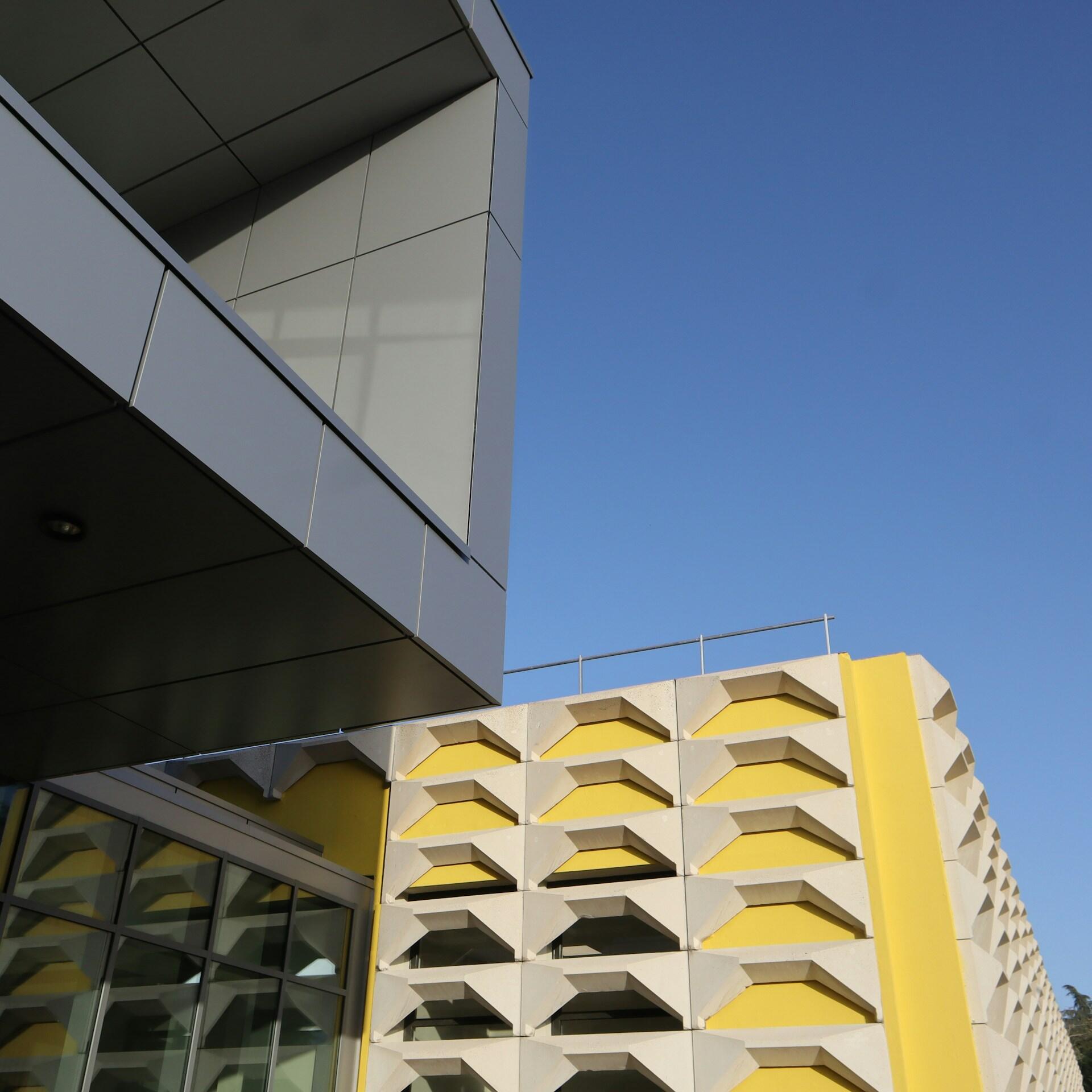 Edificio moderno de color amarillo y blanco con balcones angulosos y una entrada acristalada, visto desde abajo contra un cielo azul claro; arquitectura geométrica llamativa.
