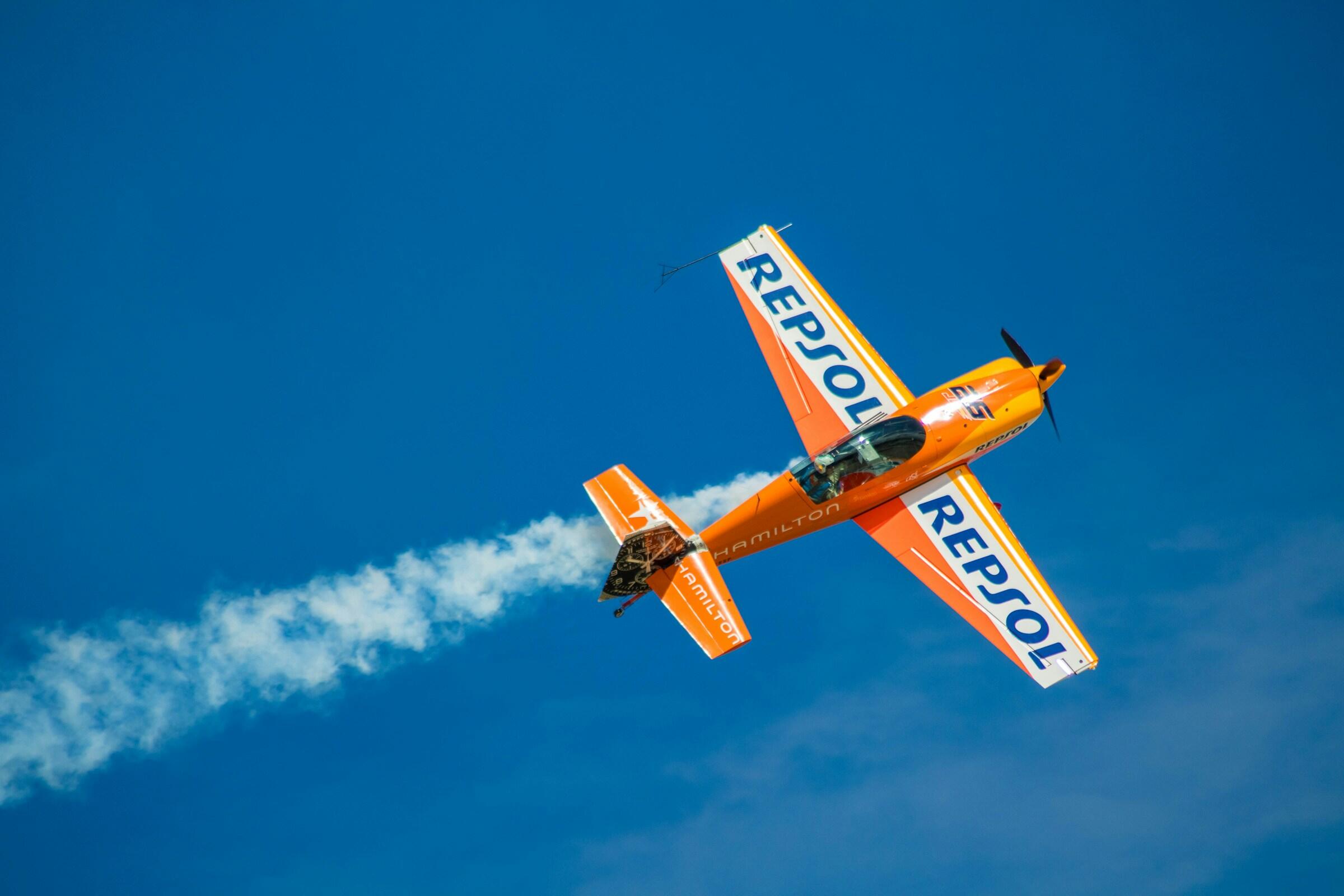 Un avión acrobático naranja con la marca «Repsol» realiza una acrobacia, dejando una estela de humo blanco contra un cielo azul claro.