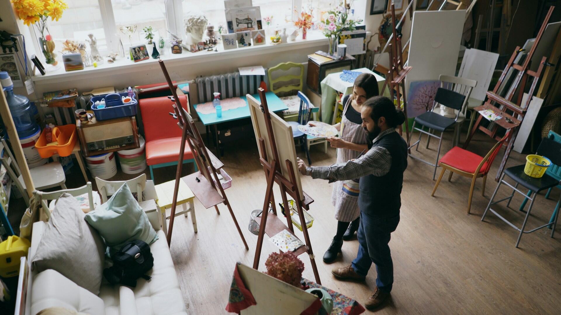 Dos artistas pintan frente a sus caballetes en un estudio luminoso y acogedor, lleno de lienzos, sillas, plantas y materiales artísticos, lo que crea un ambiente de concentración.