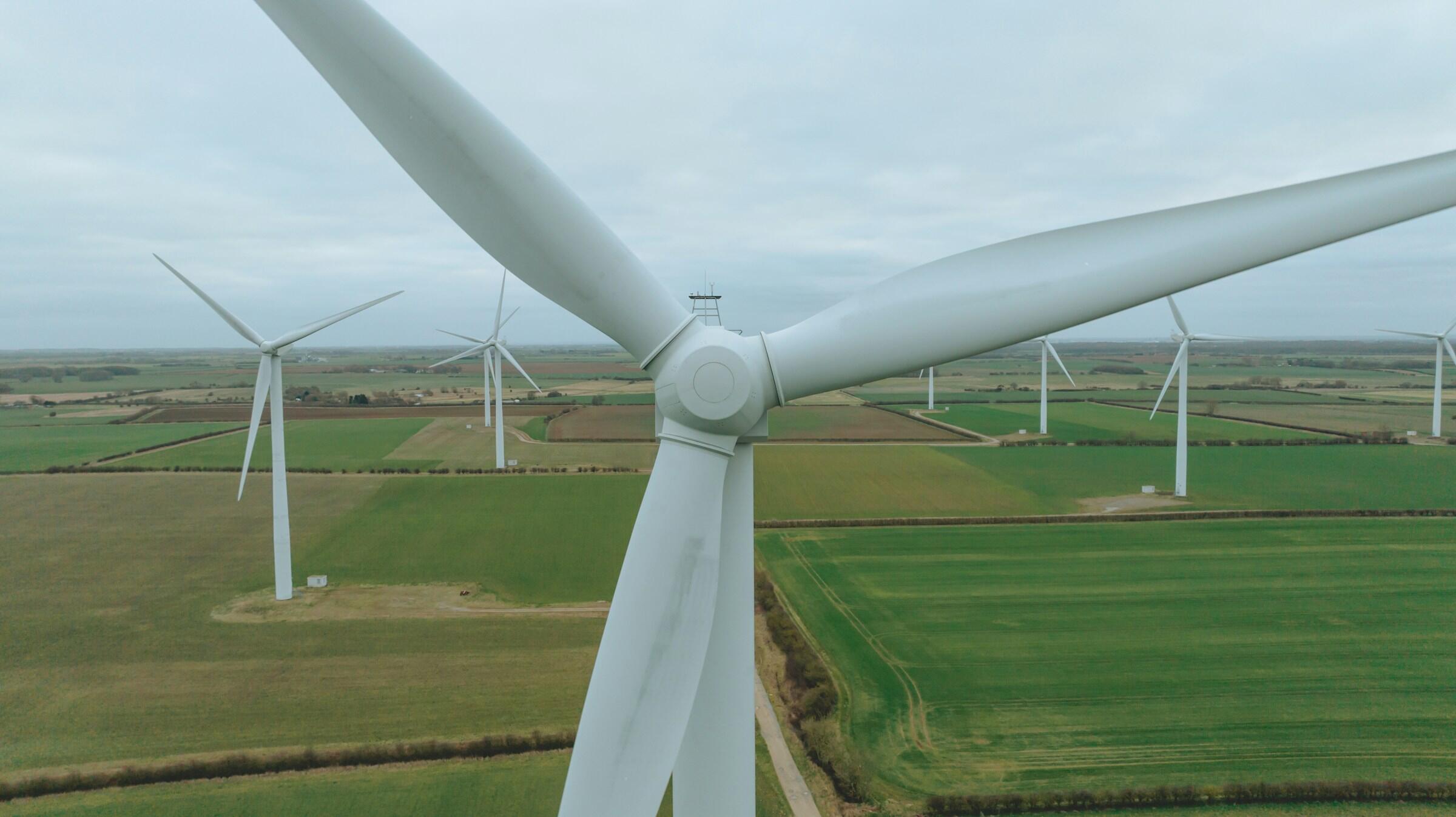 Primer plano de una moderna turbina eólica con palas giratorias, con vistas a un vasto paisaje verde salpicado de más turbinas bajo un cielo nublado.