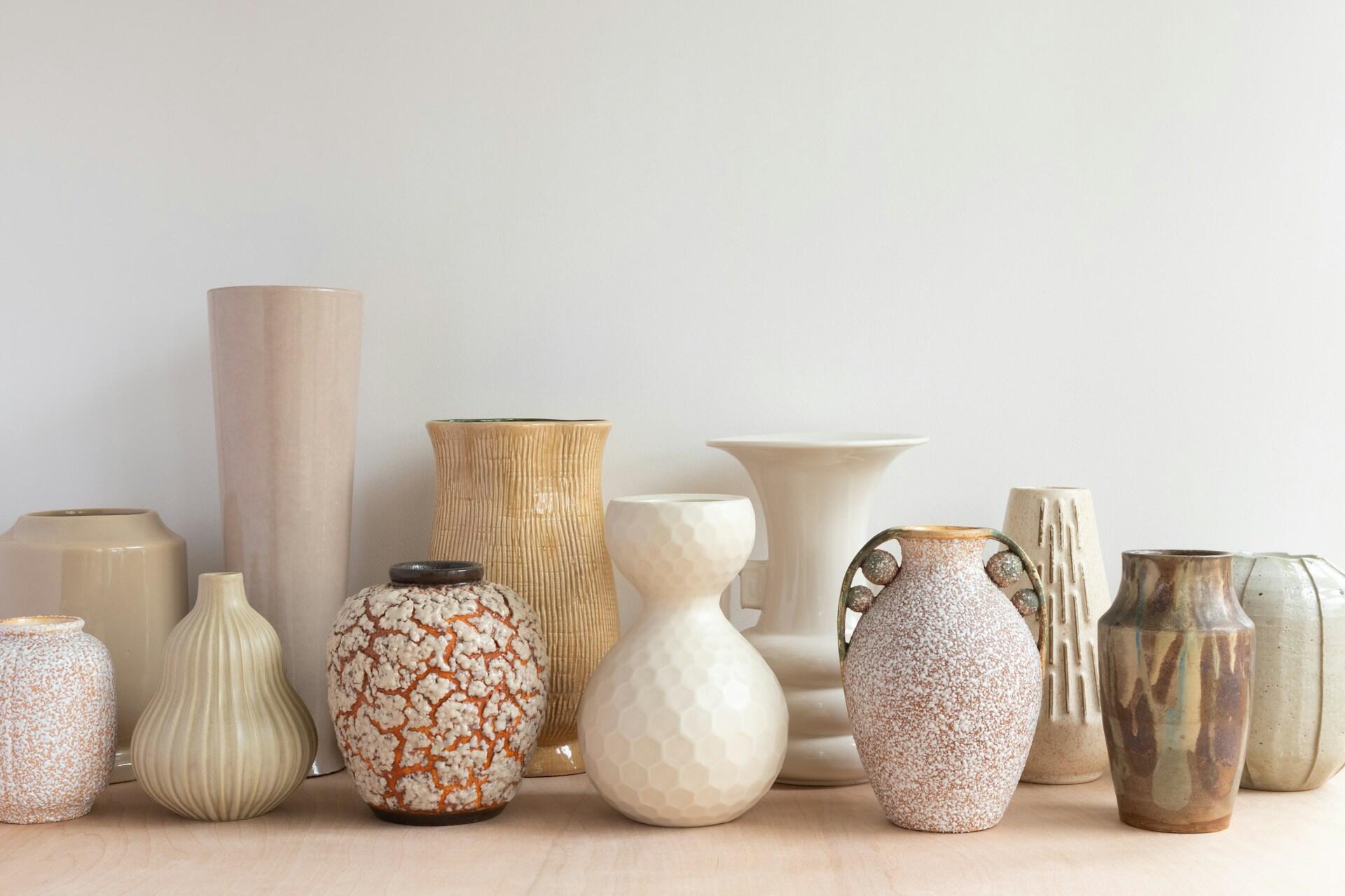 Una colección de jarrones decorativos de diversos tamaños, formas y colores neutros, dispuestos sobre una superficie de madera con un fondo blanco.