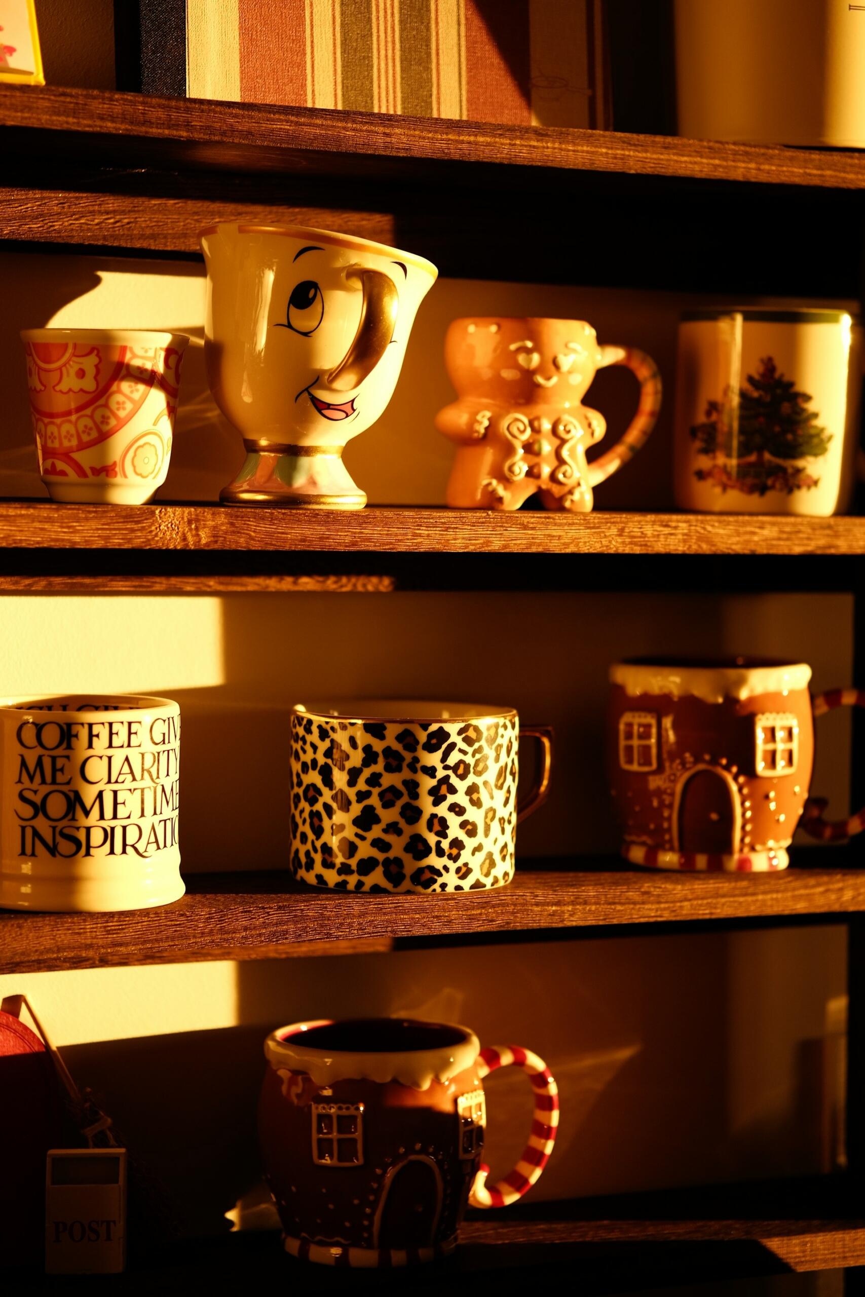 Una estantería de madera muestra una variedad de tazas de colores, entre las que se incluyen una taza con una sonrisa, una taza con un hombrecito de jengibre y una con un diseño de estampado de leopardo, bañadas por la cálida luz del sol.
