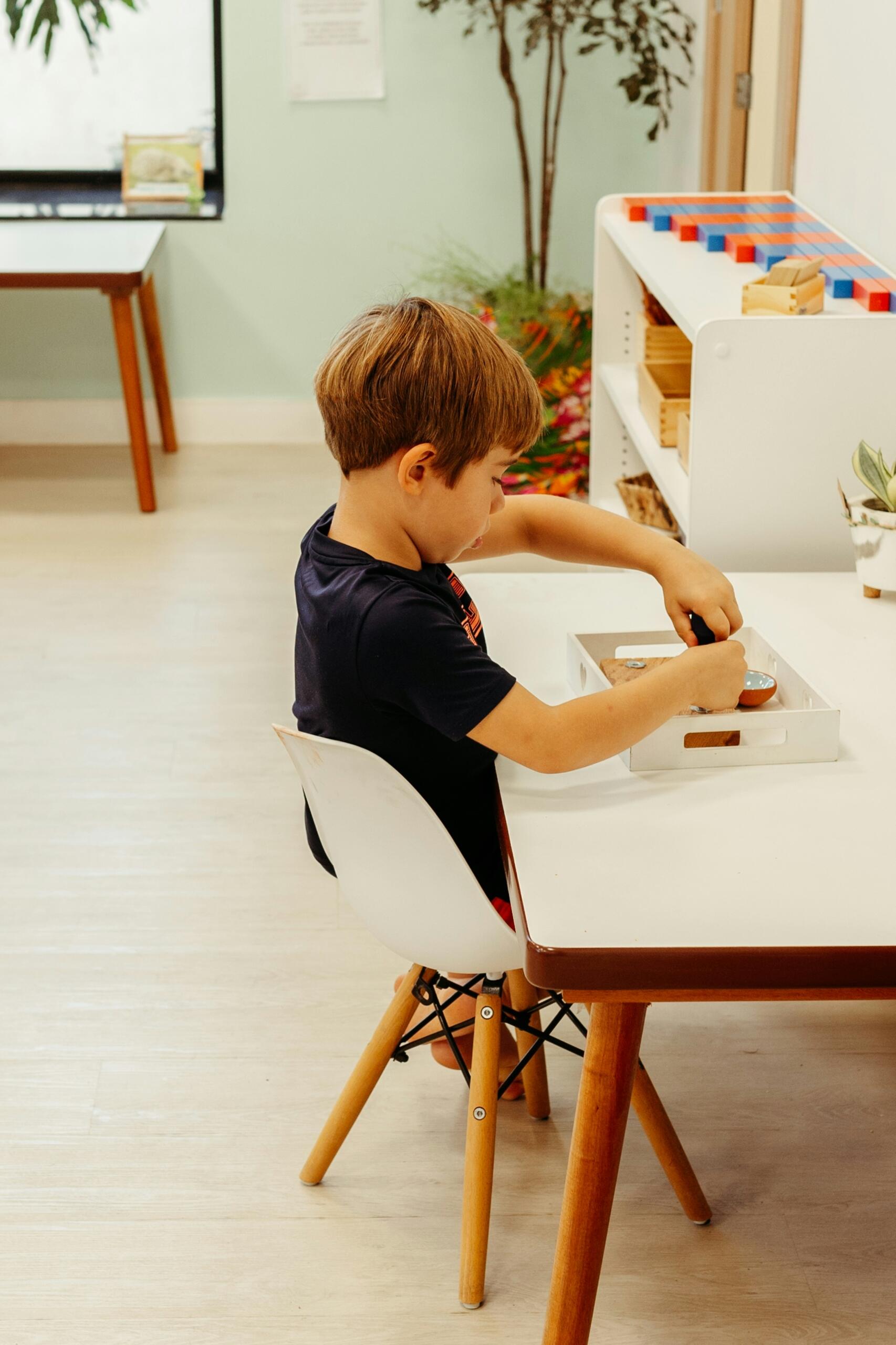 Un niño está sentado en una mesa blanca, concentrado en una actividad con una bandeja de madera llena de herramientas, en un entorno de aprendizaje luminoso y colorido.