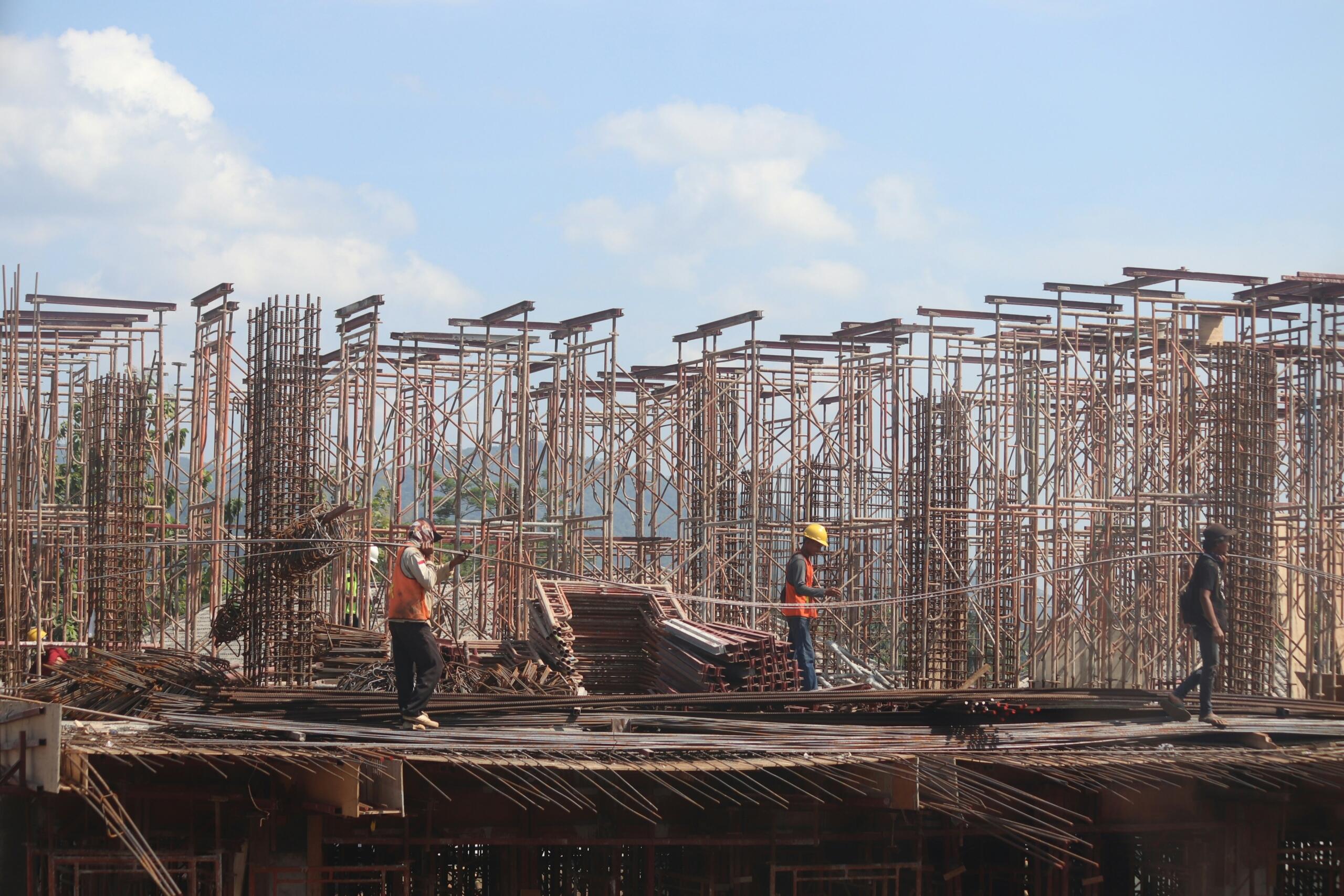 Se ve a unos trabajadores de la construcción en una obra parcialmente terminada, rodeados de estructuras de acero y un cielo azul brillante.