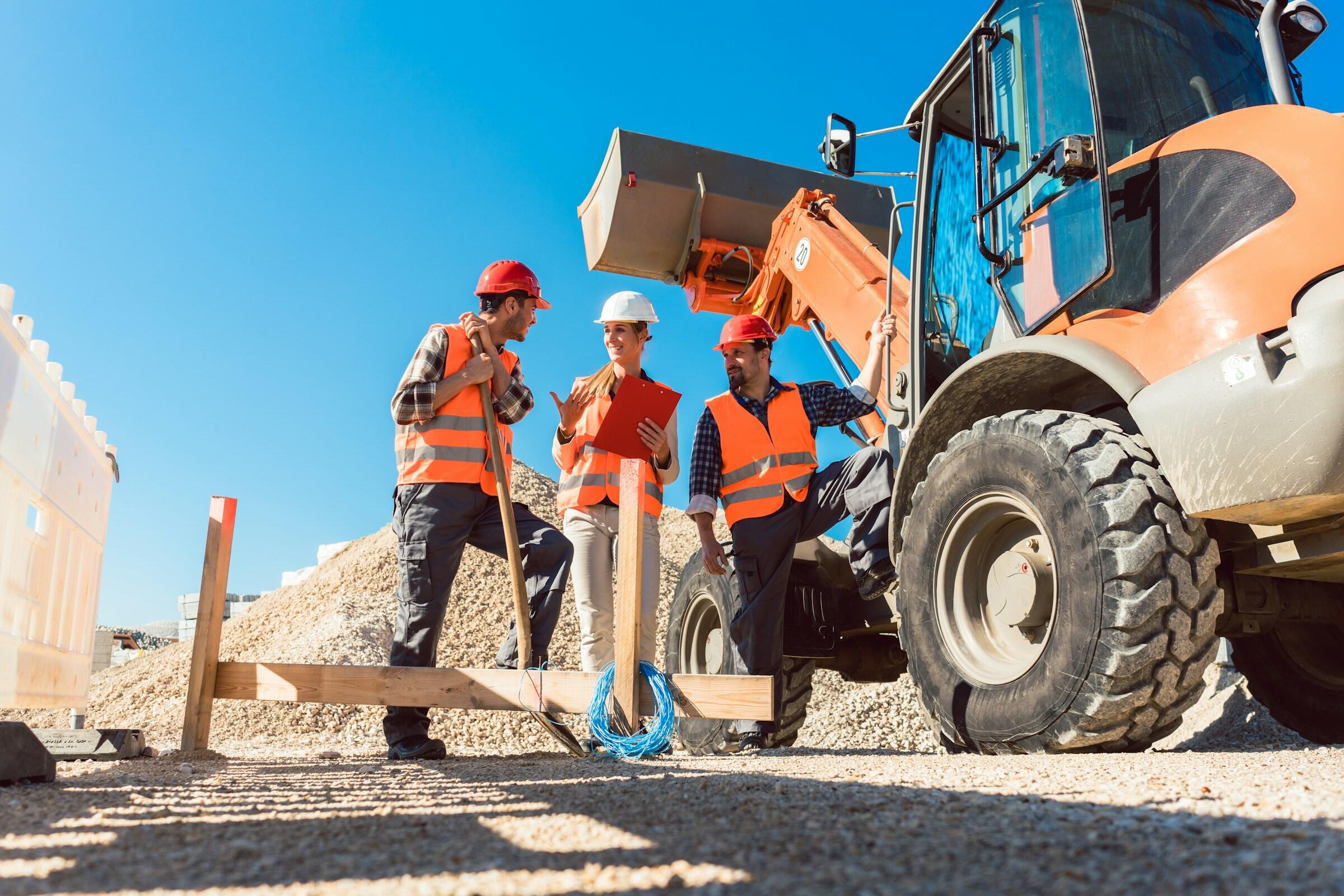 Los trabajadores de la construcción, equipados con chalecos de seguridad y cascos, discuten los planes cerca de la maquinaria pesada en un día soleado en la obra.