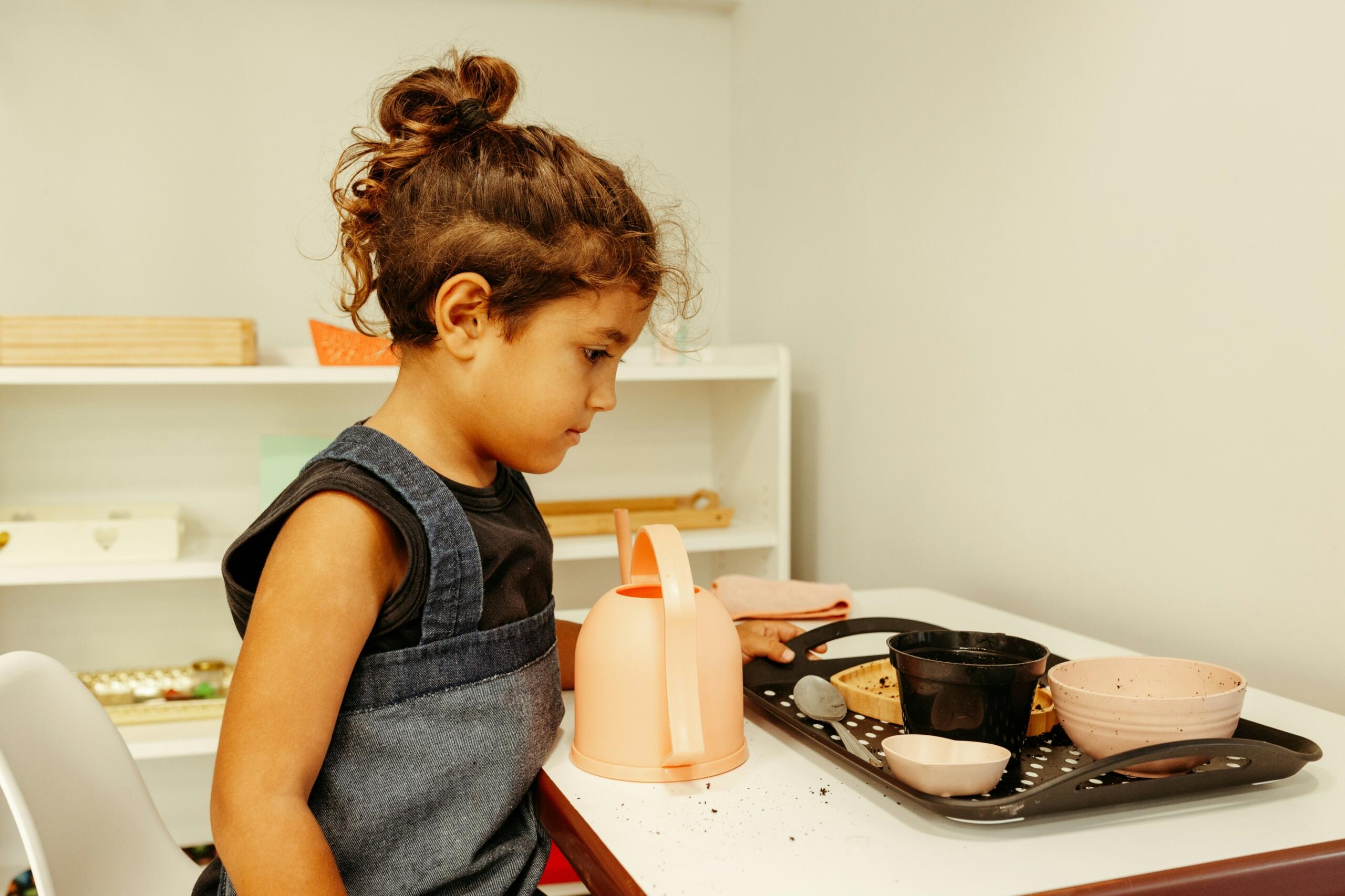 Una niña o  niño con el pelo rizado está de pie junto a una bandeja llena de ollas, una tetera y utensilios en una sala de juegos luminosa y organizada.
