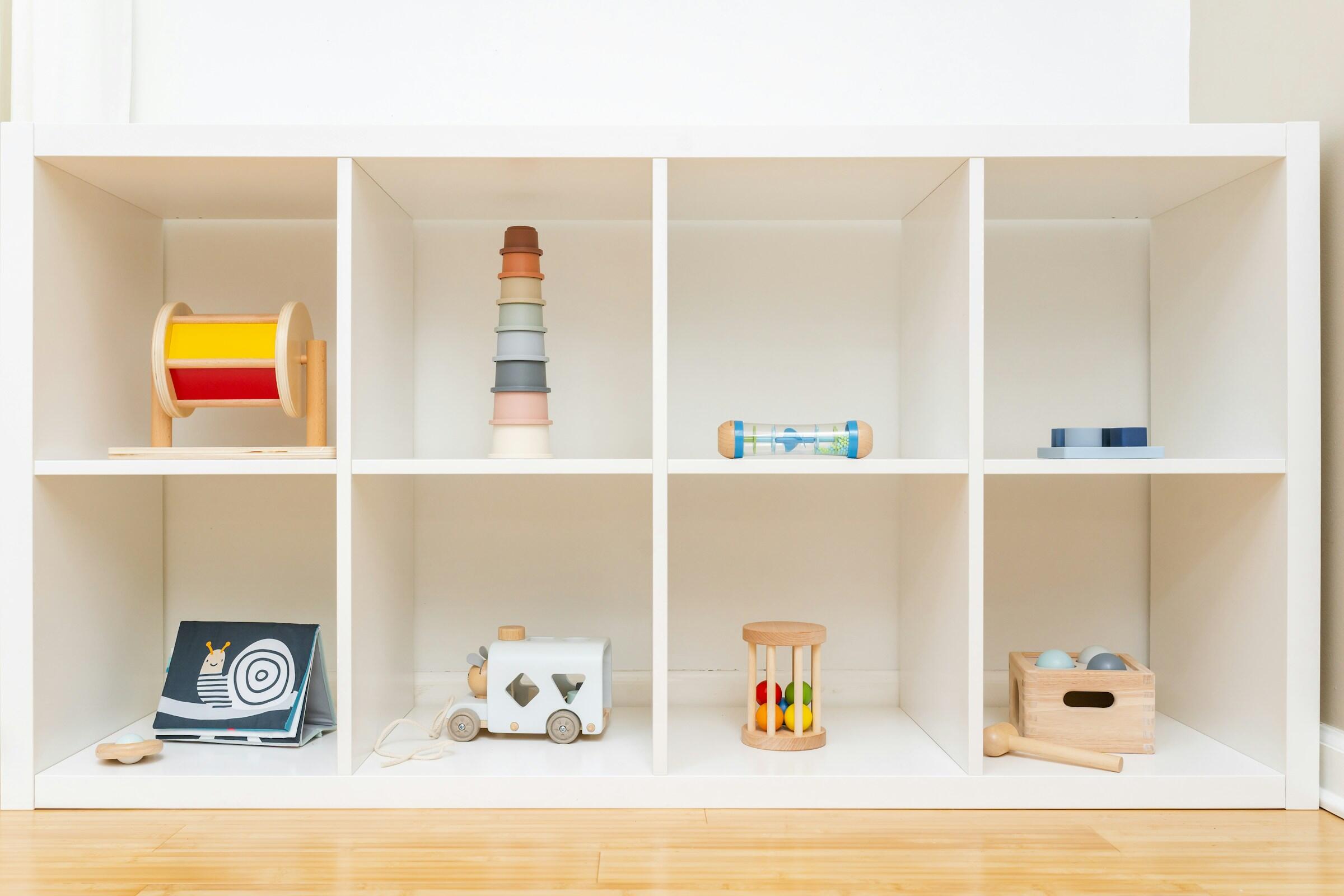 Juguetes de madera de colores vivos cuidadosamente dispuestos en una estantería blanca, entre los que se incluyen un tambor, vasos apilables y un tren de juguete.