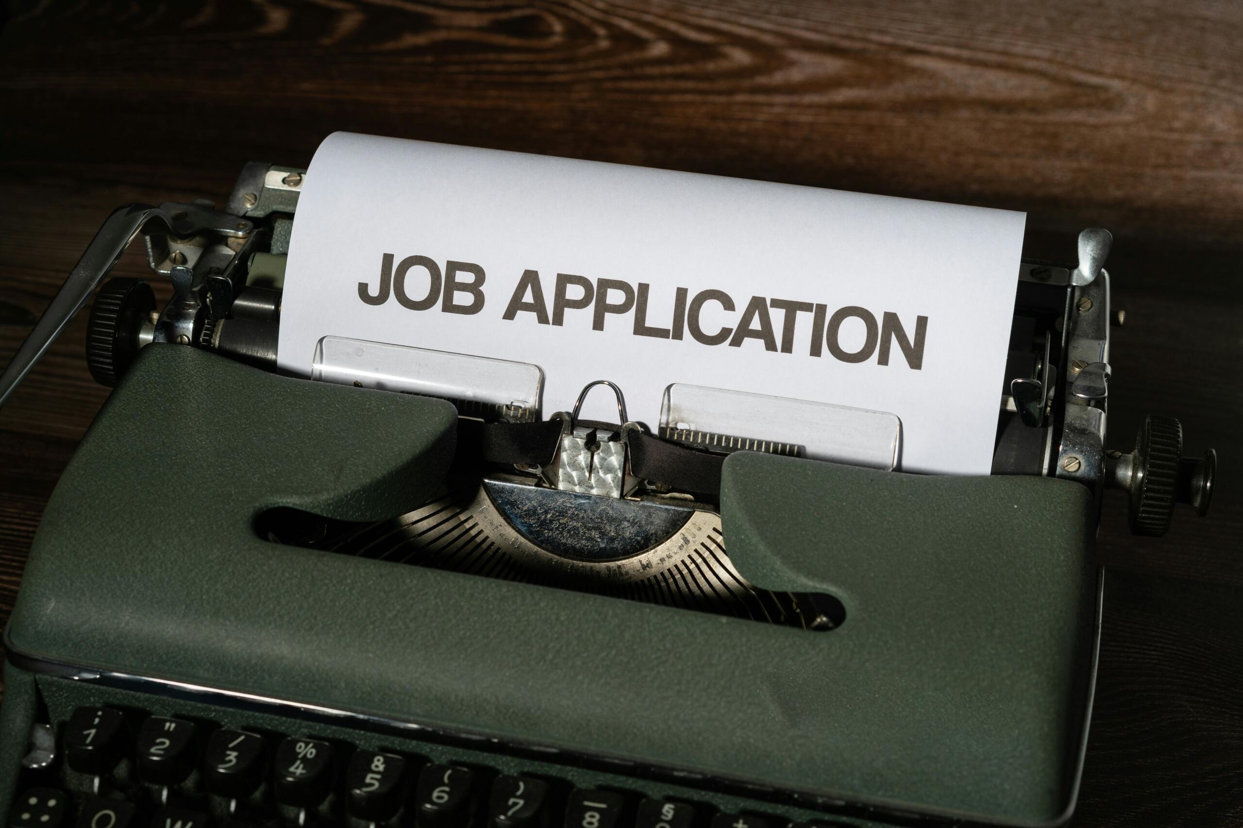 Una máquina de escribir verde vintage con una hoja de papel blanco en la que se lee «SOLICITUD DE EMPLEO» en negrita.