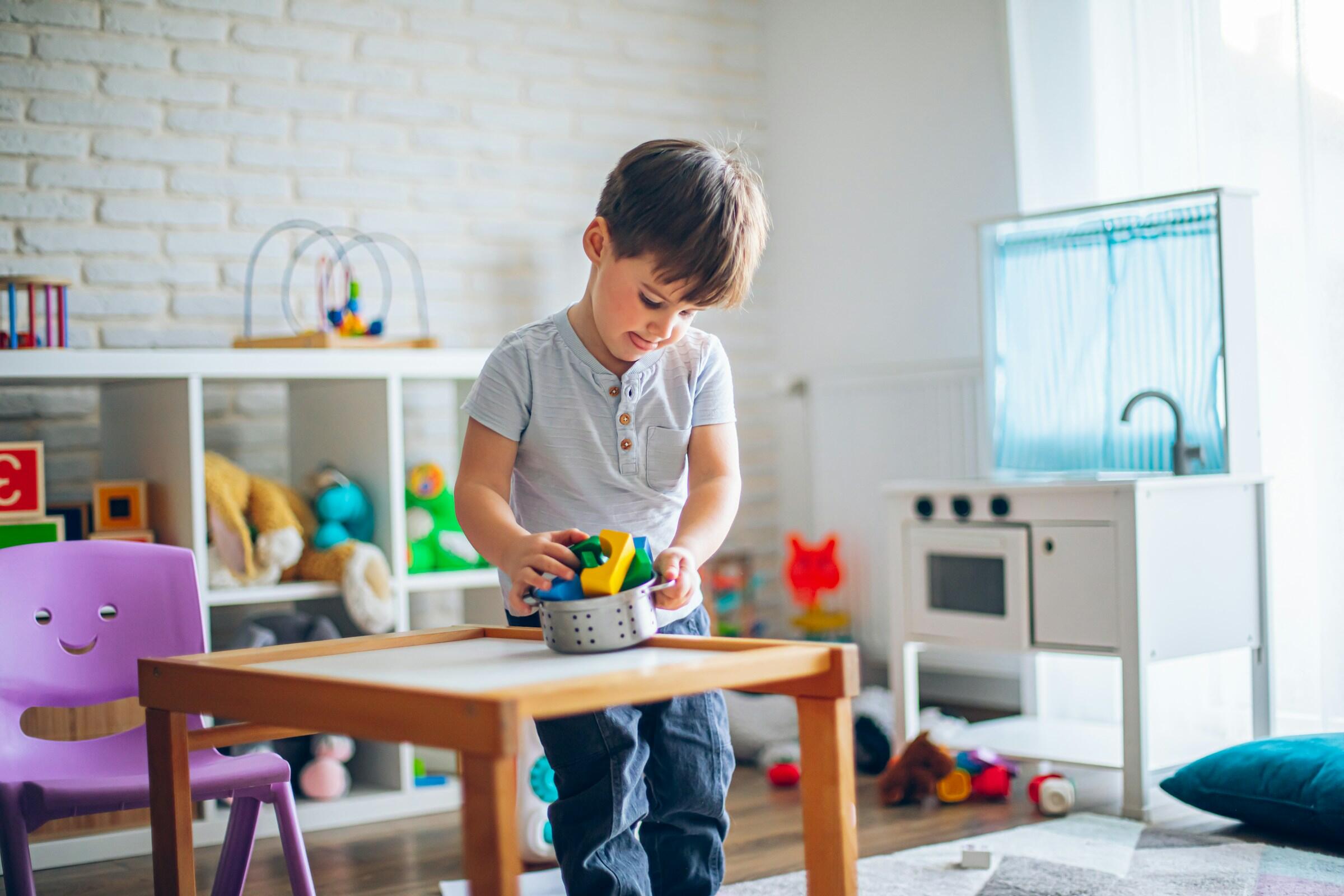 Un niño en una habitación luminosa juega en una mesa de madera, organizando bloques de construcción de colores que sacó de un cuenco plateado.