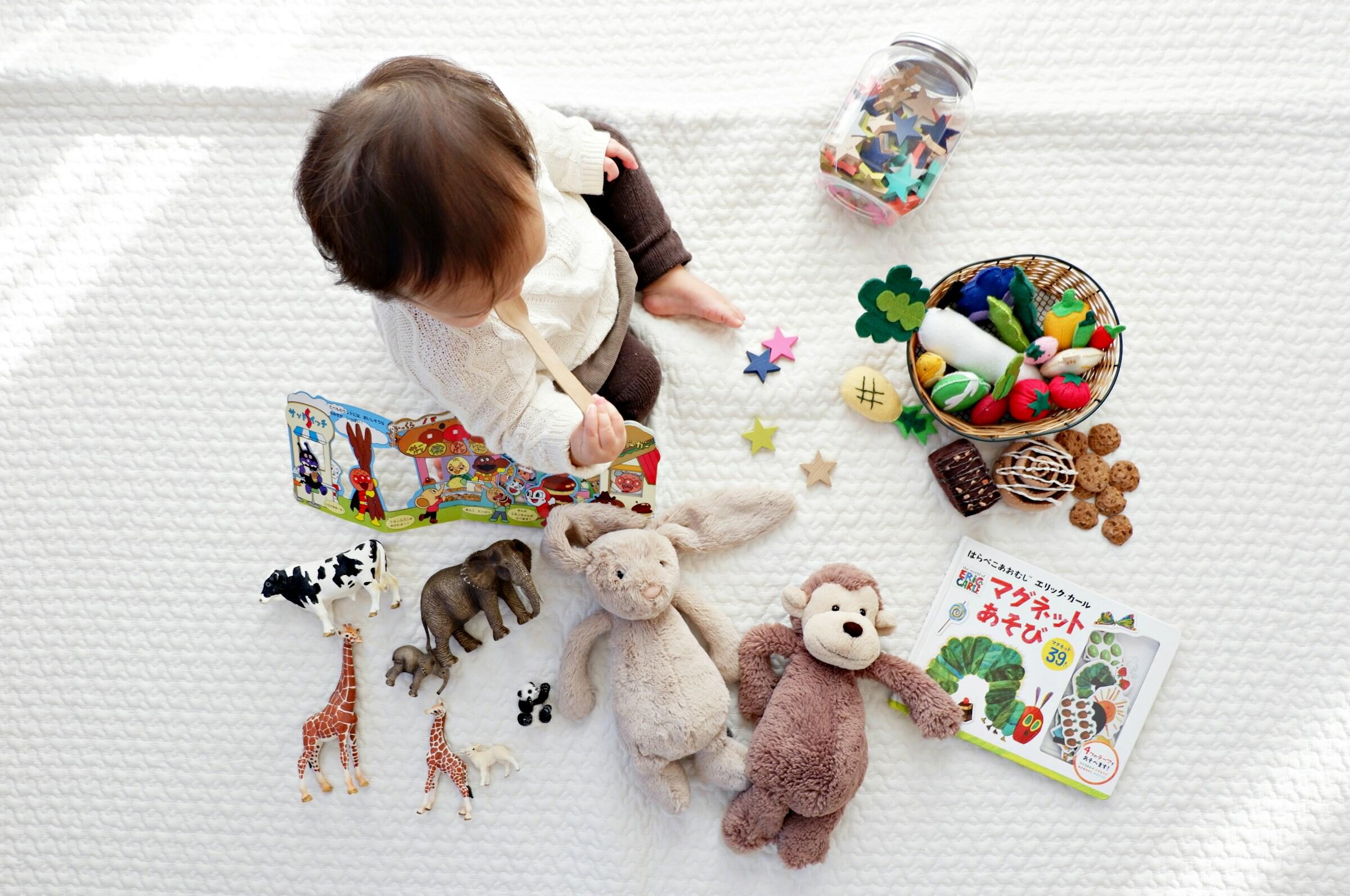 Un niño pequeño sentado sobre una manta blanca juega con un libro de colores, rodeado de peluches, formas divertidas y figuras de animales variadas.