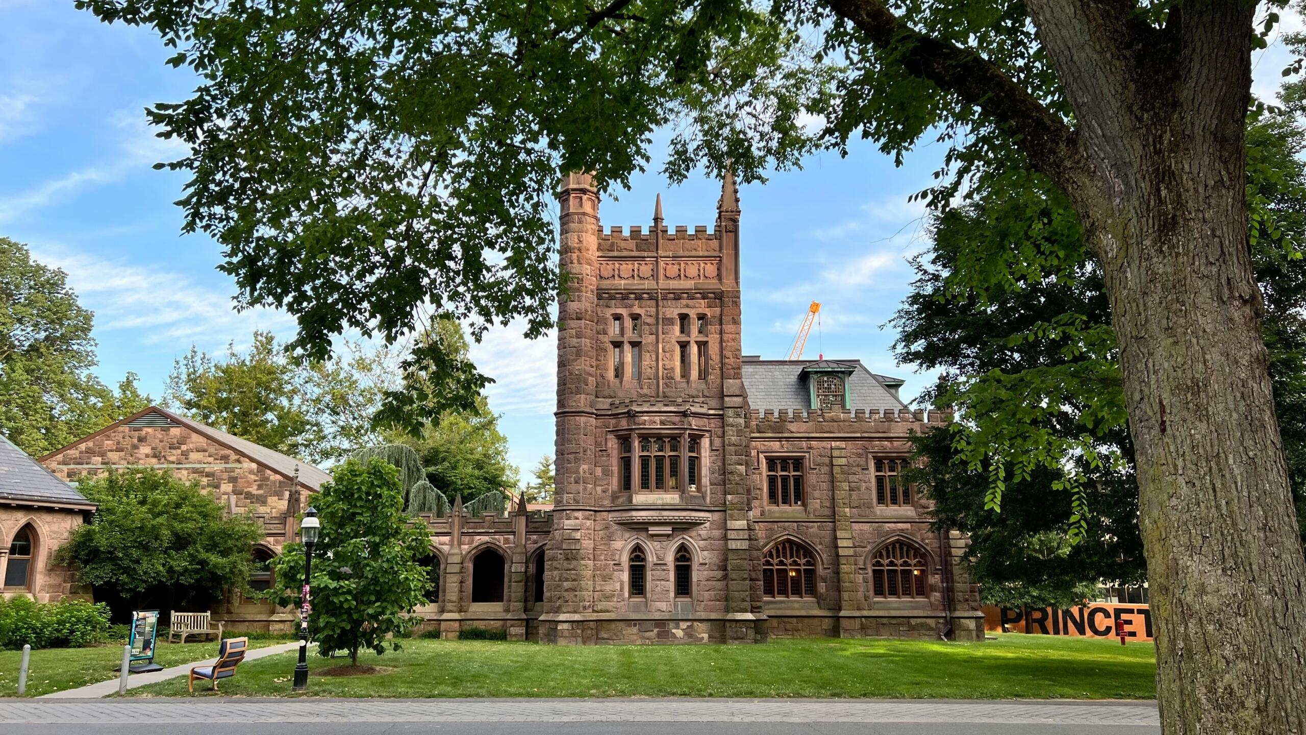 Edificio histórico de piedra con torres ornamentadas, rodeado de exuberante vegetación y con un letrero que dice Princeton.