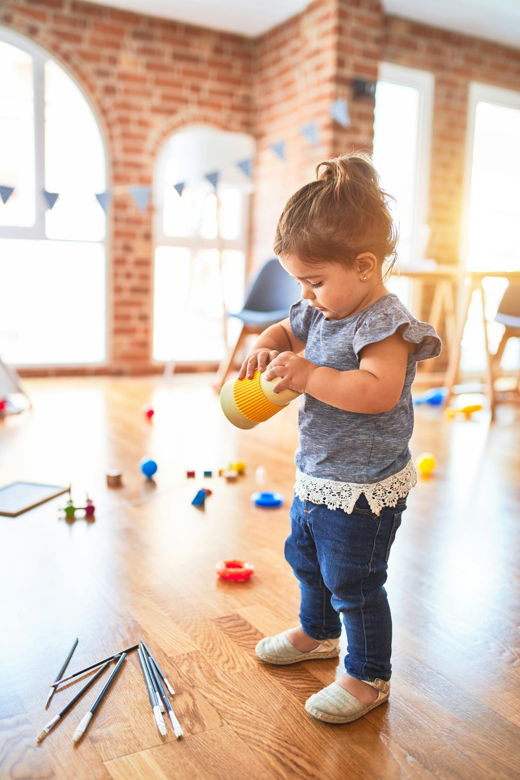 Una niña pequeña vestida con una camiseta azul y vaqueros está de pie sobre un suelo de madera, sosteniendo una taza amarilla, rodeada de juguetes de colores esparcidos por el suelo.