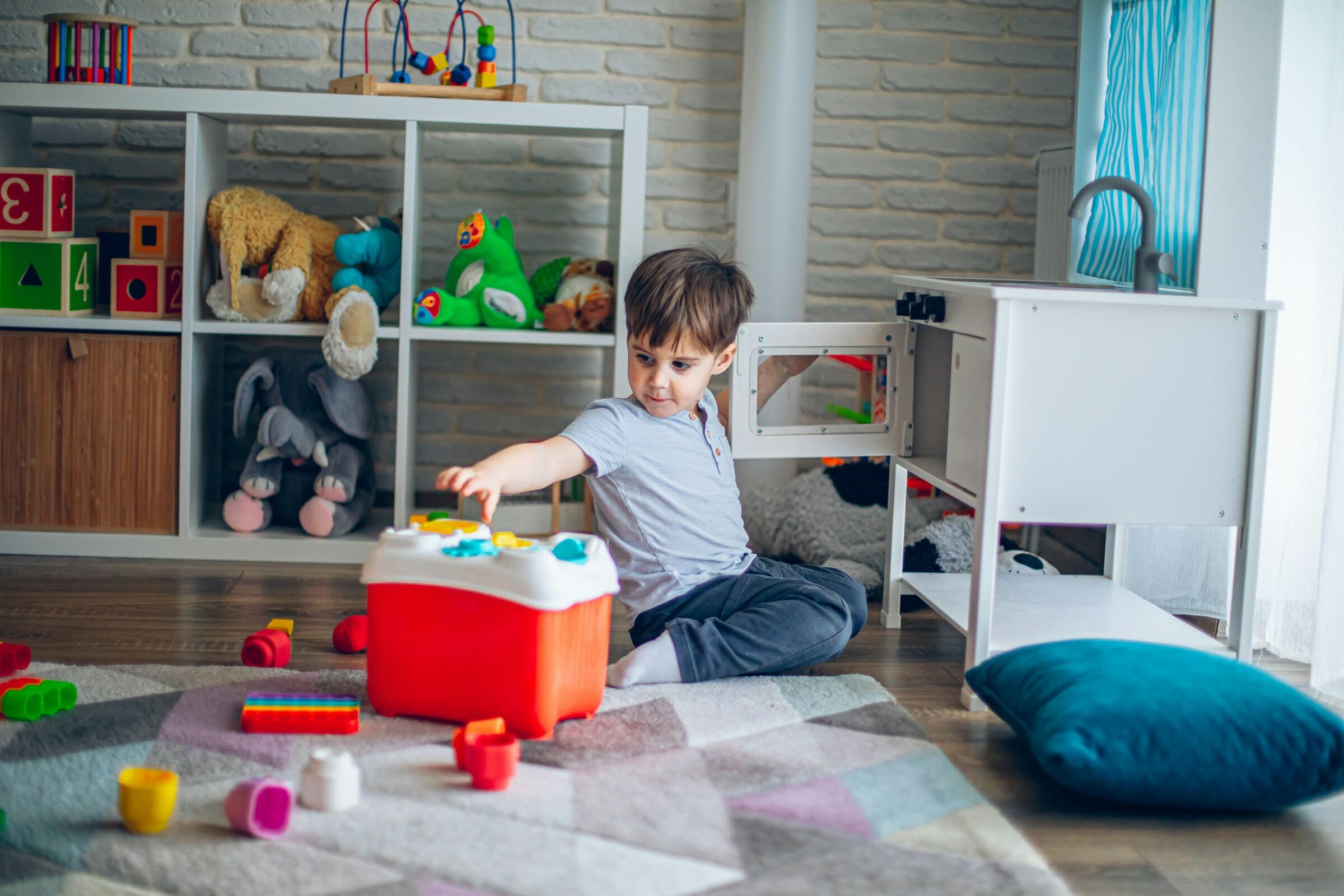Un niño está sentado sobre una alfombra de colores rodeado de juguetes, jugando con un baúl rojo en una sala de juegos luminosa y acogedora.